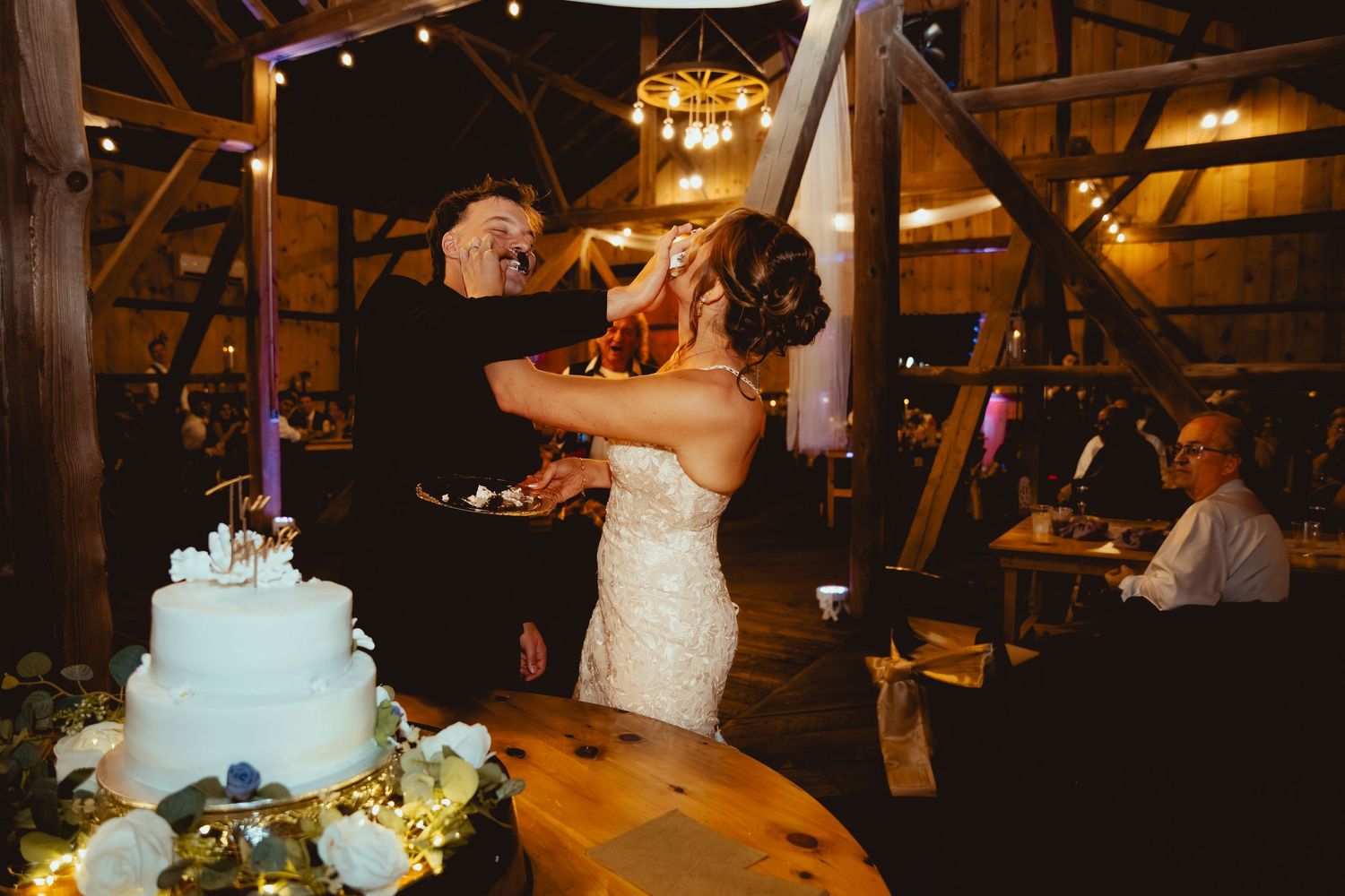 Wedding reception in a dimly lit rustic barn venue with string lights and elegant white wedding cake display.