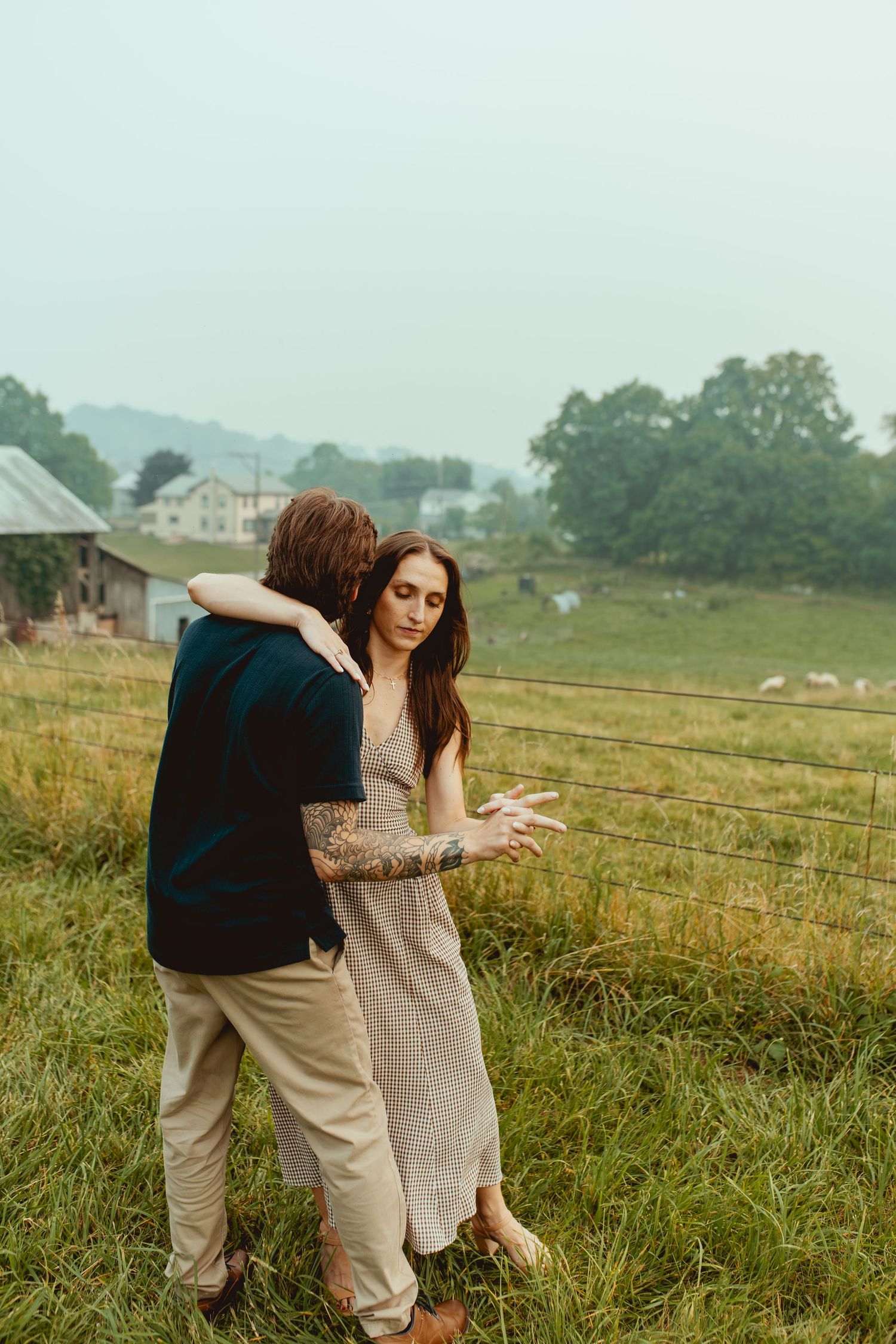 A couple embraces in a green field, woman in floral dress, tattooed man in dark jacket, rural farm backdrop.