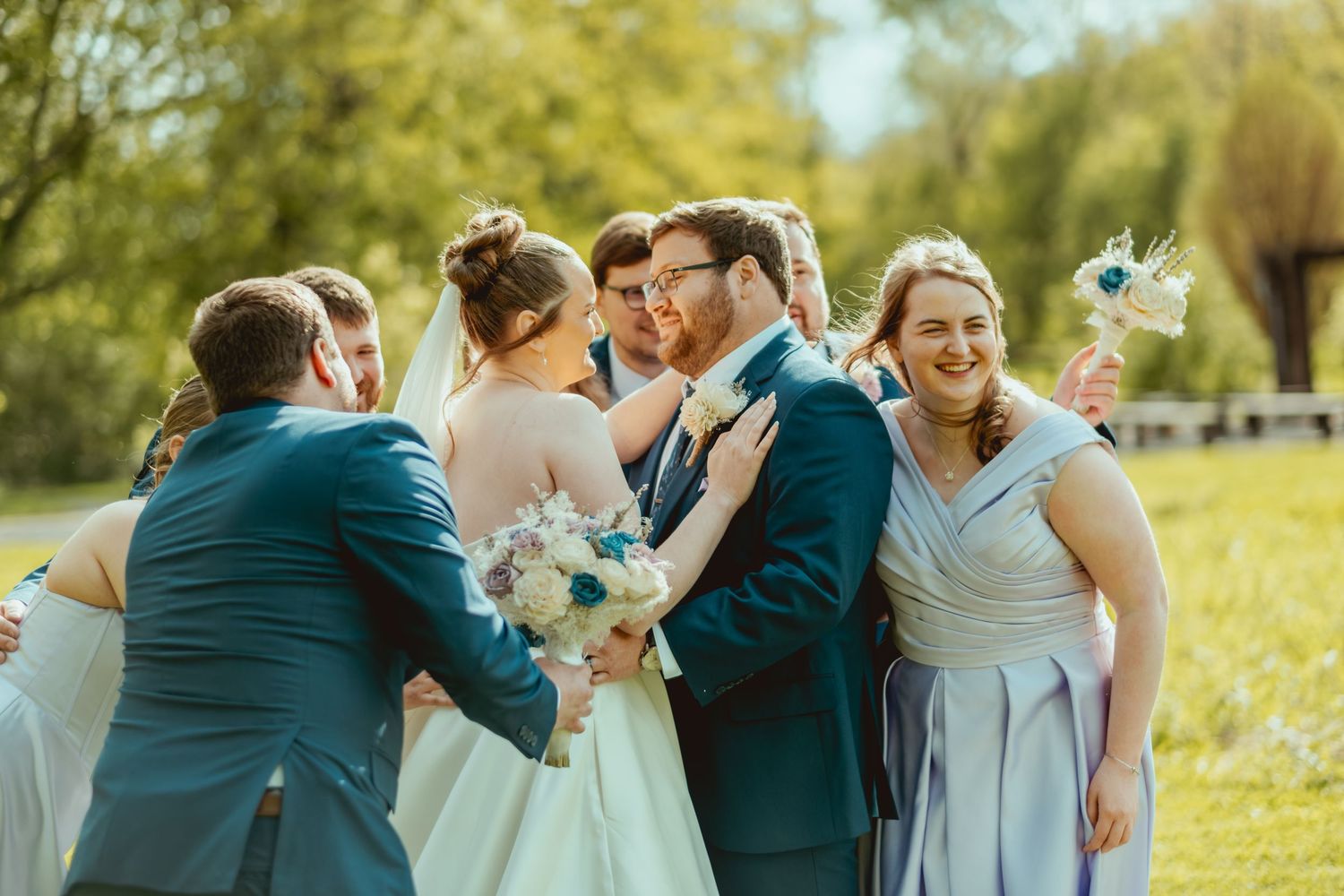 Wedding party surrounds smiling bride and groom outdoors, bridesmaids in blue dresses holding bouquets on sunny day.