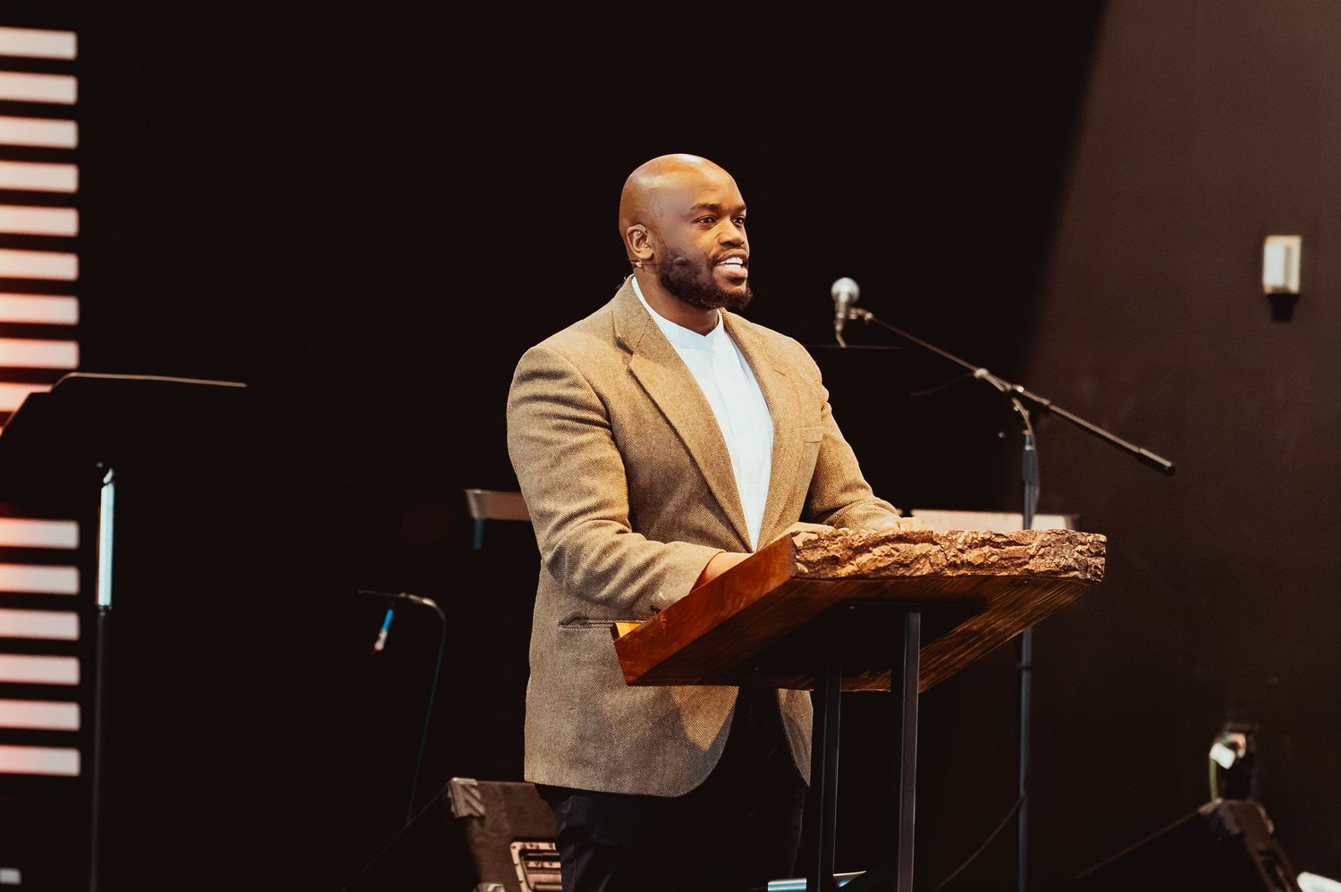 A speaker in a tan blazer and light blue shirt delivers remarks from a wooden podium on stage.