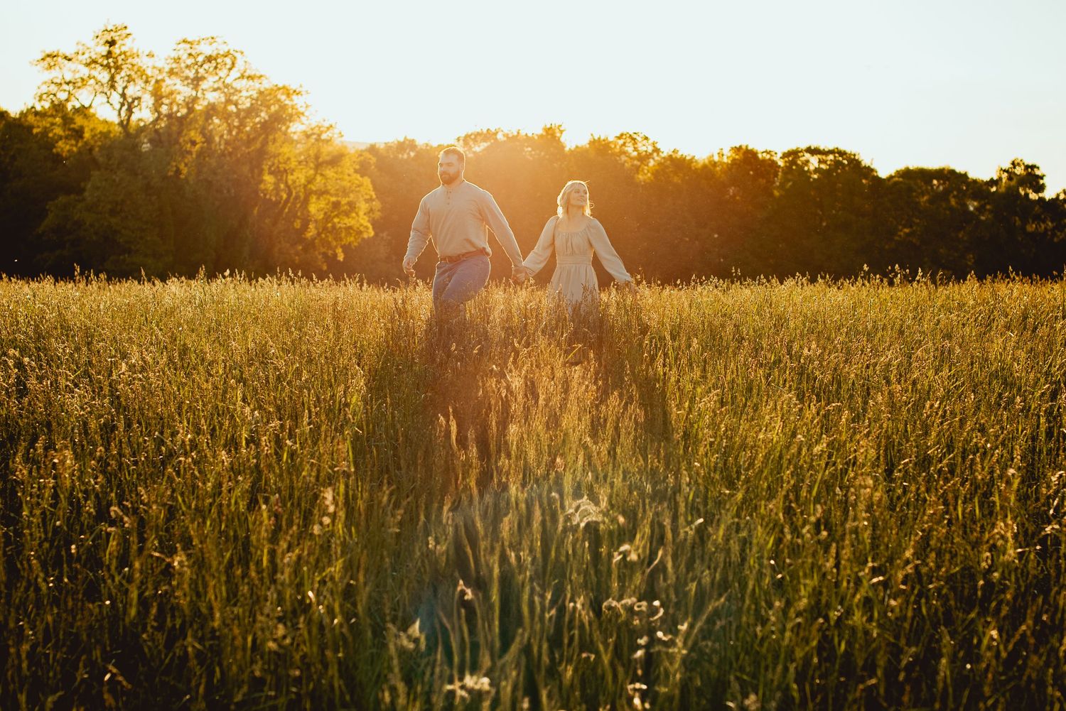 Two people holding hands while walking through a golden meadow at sunset with tall grass and wildflowers surrounding them.