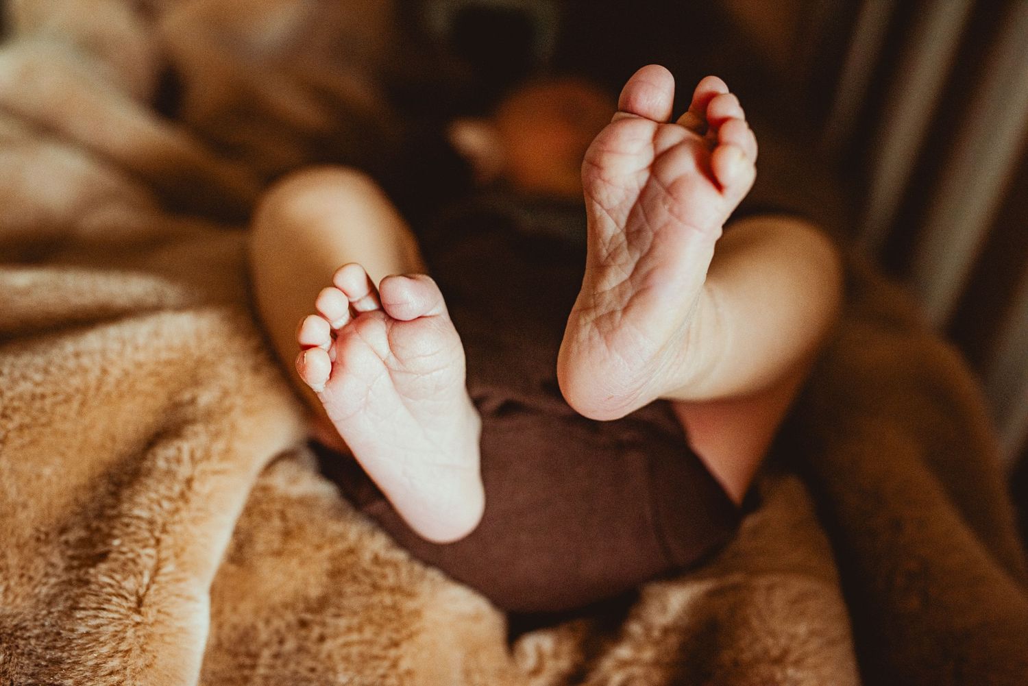 A tiny newborn baby's feet being gently held against a warm brown blanket in natural lighting.