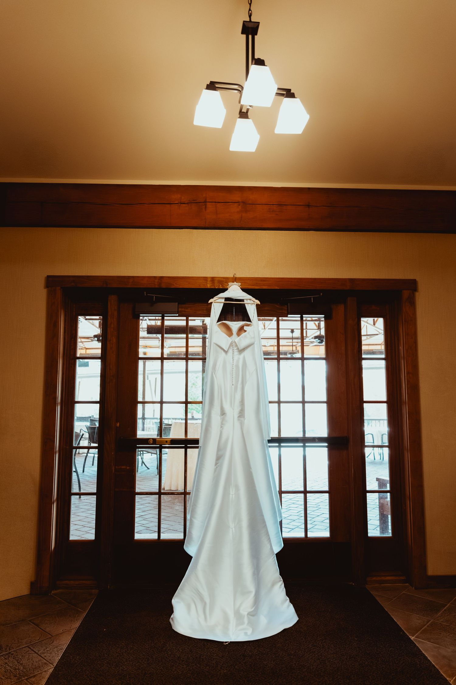 A white wedding dress hangs in silhouette against a row of dark wooden windows in a warmly lit room.