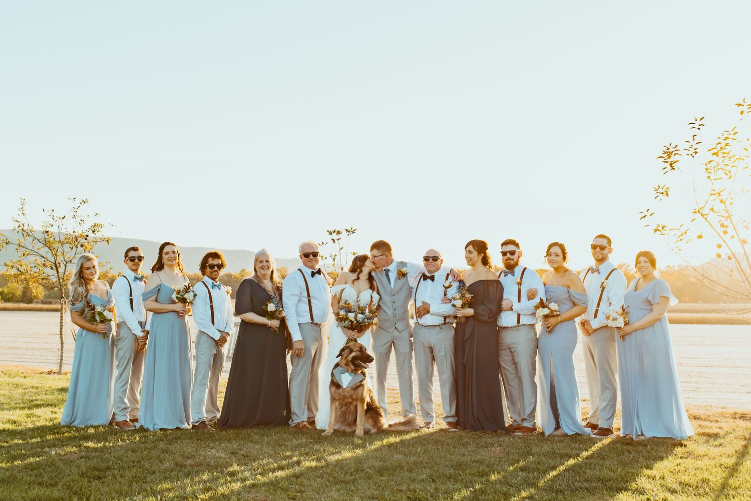Wedding party in light blue dresses and dark suits poses together at sunset in a grassy field.