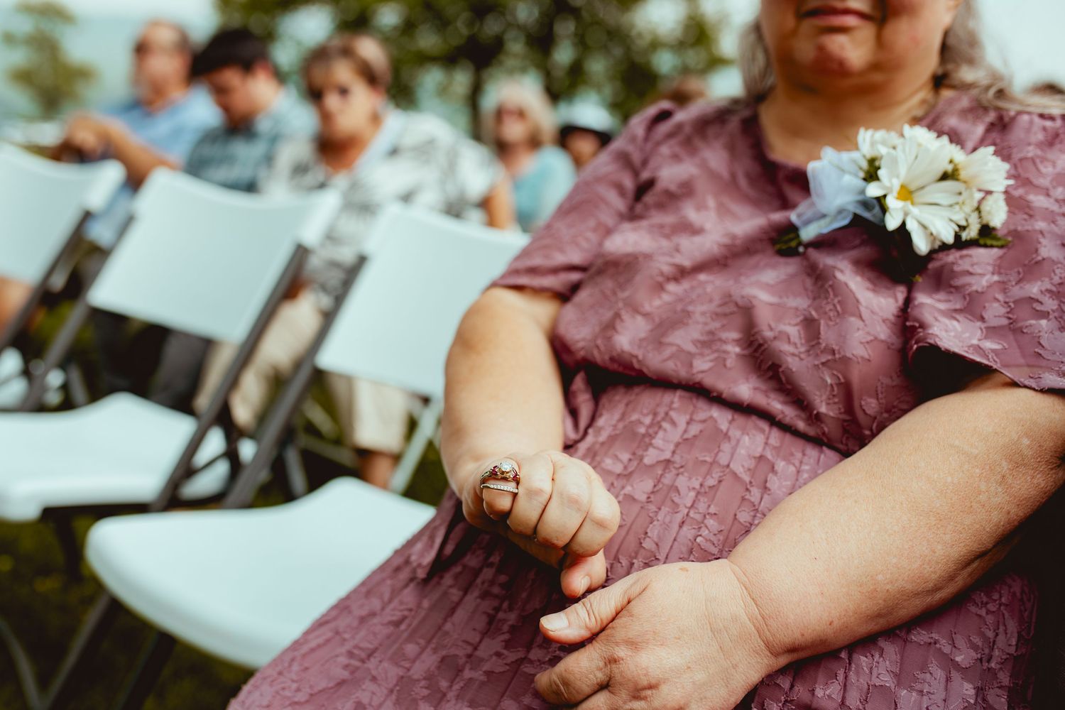Close-up of hands resting on a burgundy dress with a white corsage at an outdoor event with folding chairs in background.