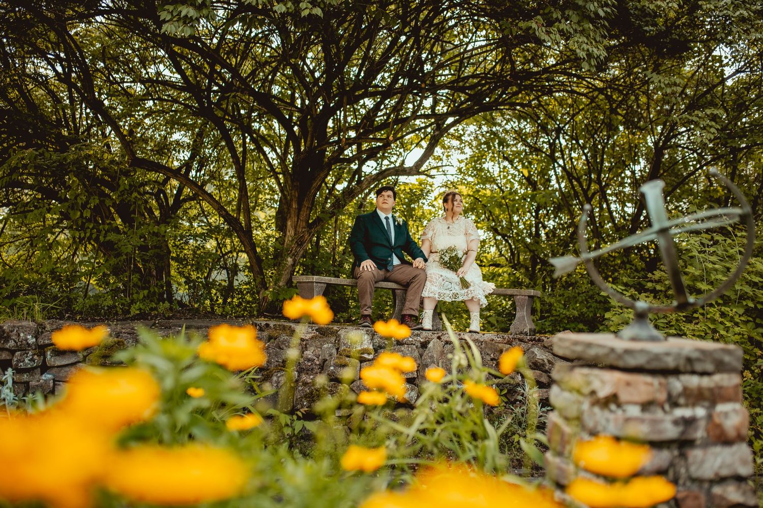 Couple walking through a rustic garden path with yellow wildflowers and stone walls in soft afternoon light.
