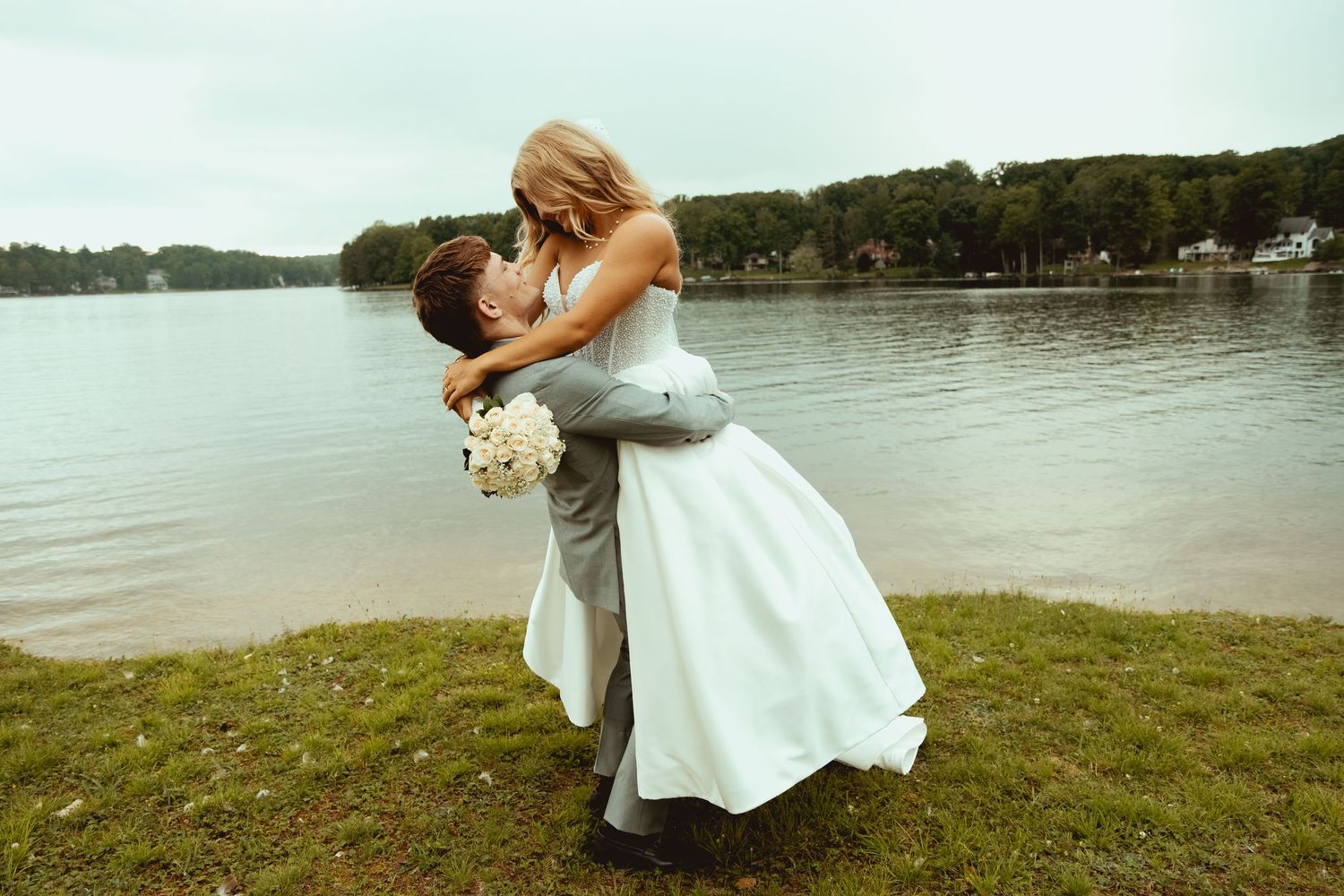 Bride in white gown lifted by groom in gray suit near lakeside, sharing romantic embrace with bouquet.