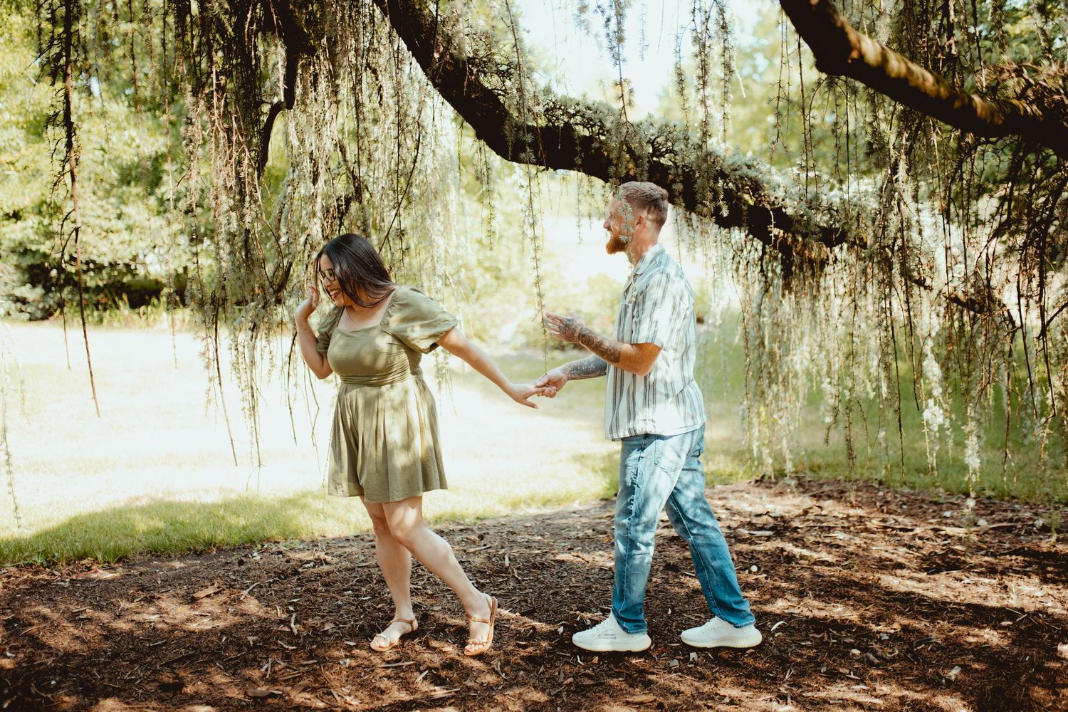 Couple dancing and holding hands under a mossy willow tree in a lush green park setting.