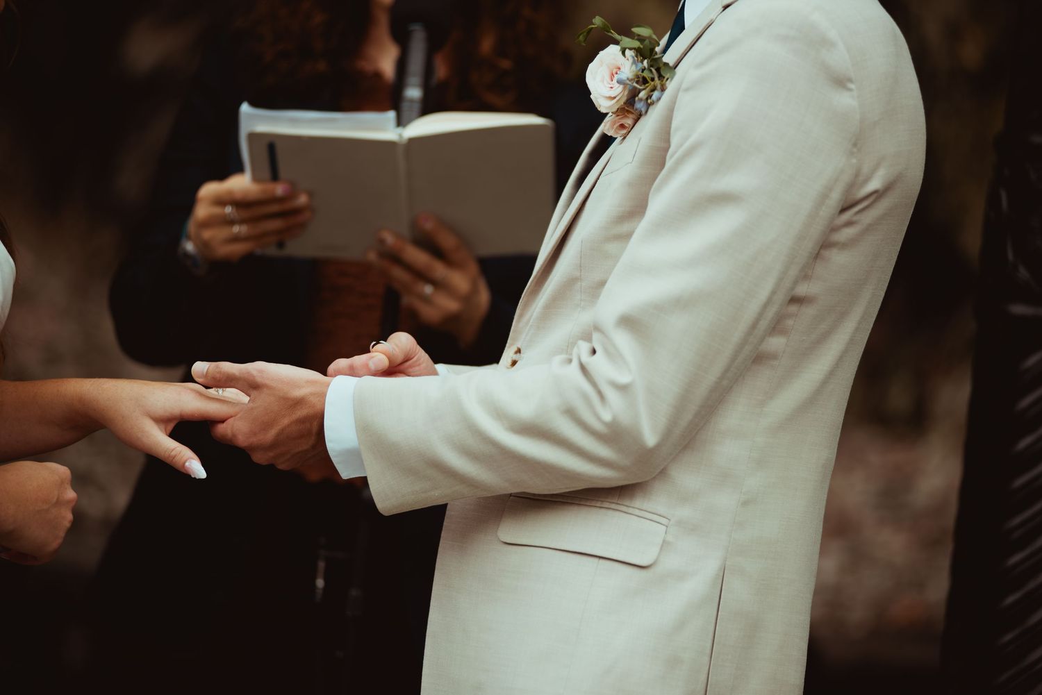 Groom in ivory suit exchanges rings with bride during outdoor wedding ceremony.