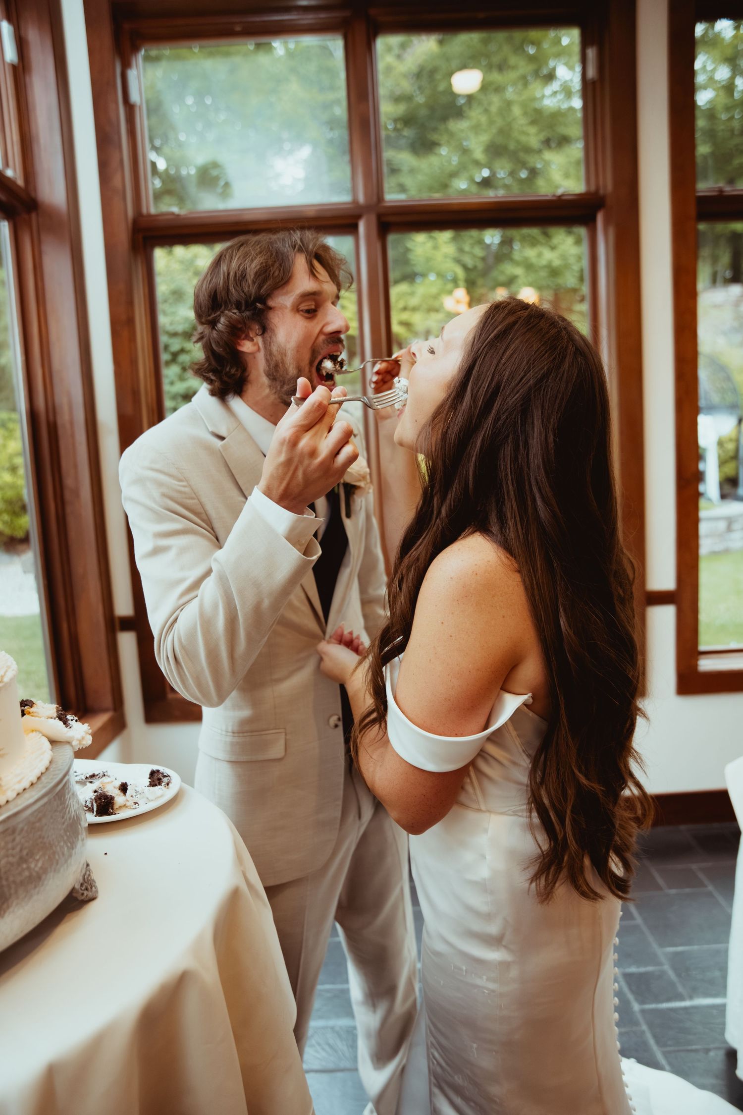Bride and groom share wedding cake cutting moment, feeding each other inside rustic reception venue.