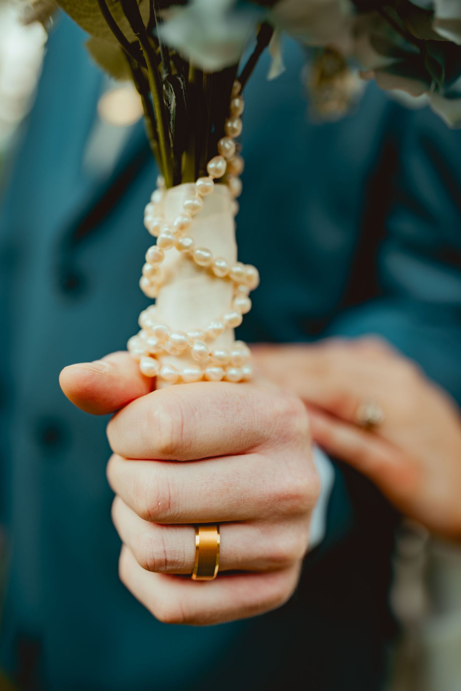 Close-up of hands holding pearl-wrapped bouquet stem with gold wedding band.