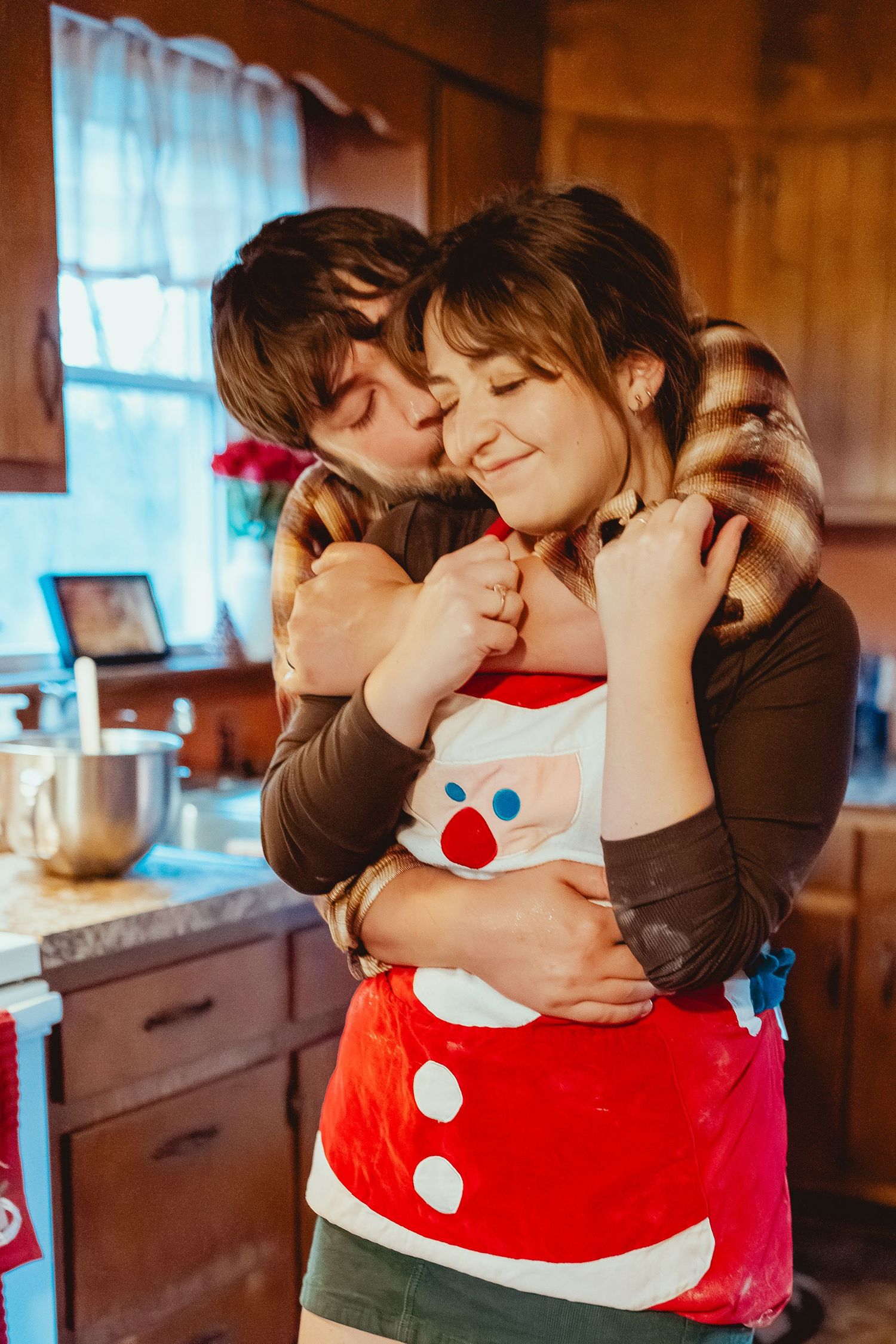Three sequential photos show a romantic couple embracing in a cozy wooden kitchen with one wearing a Santa costume.
