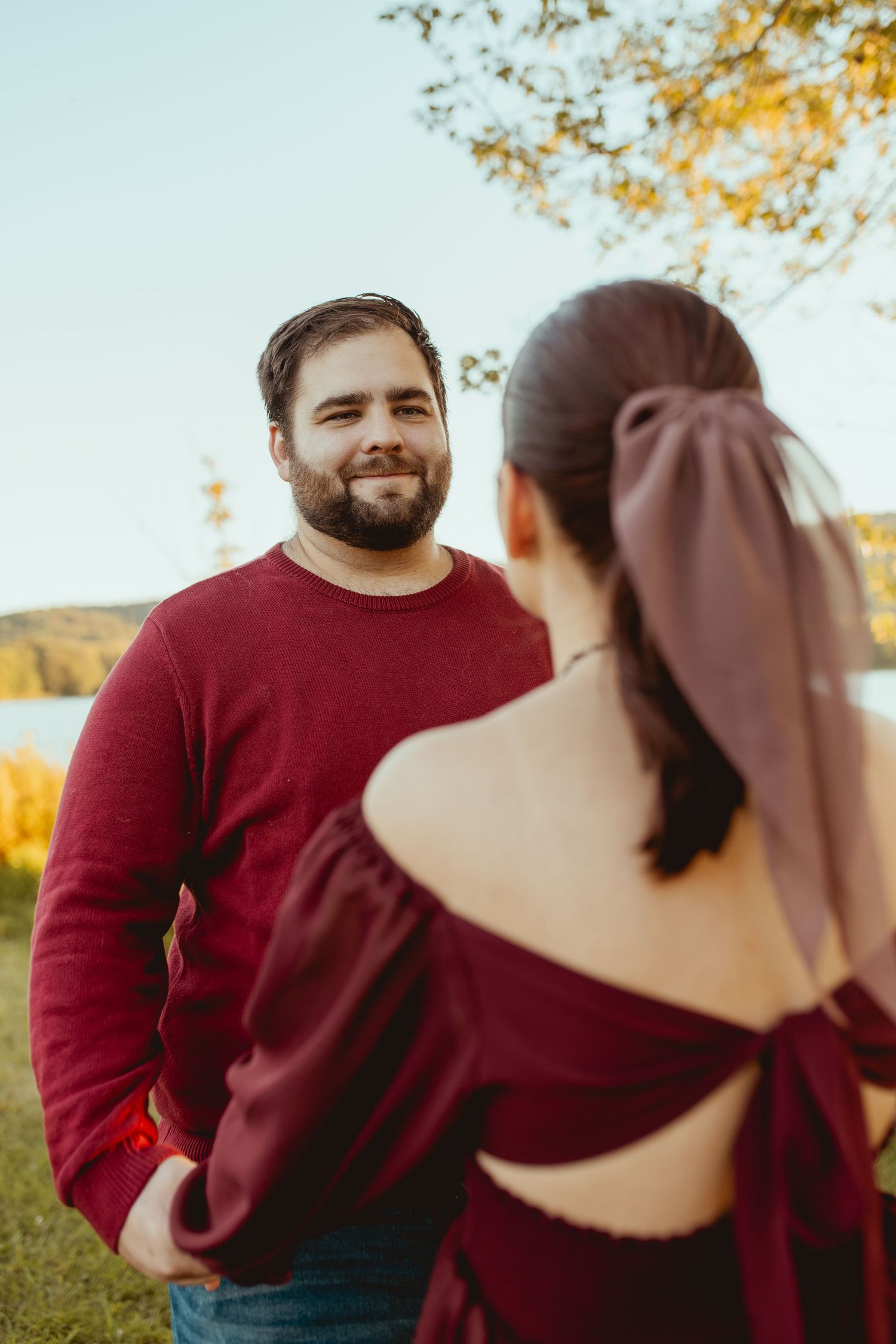 A couple in burgundy sweaters share intimate moments outdoors during a fall photoshoot at sunset.