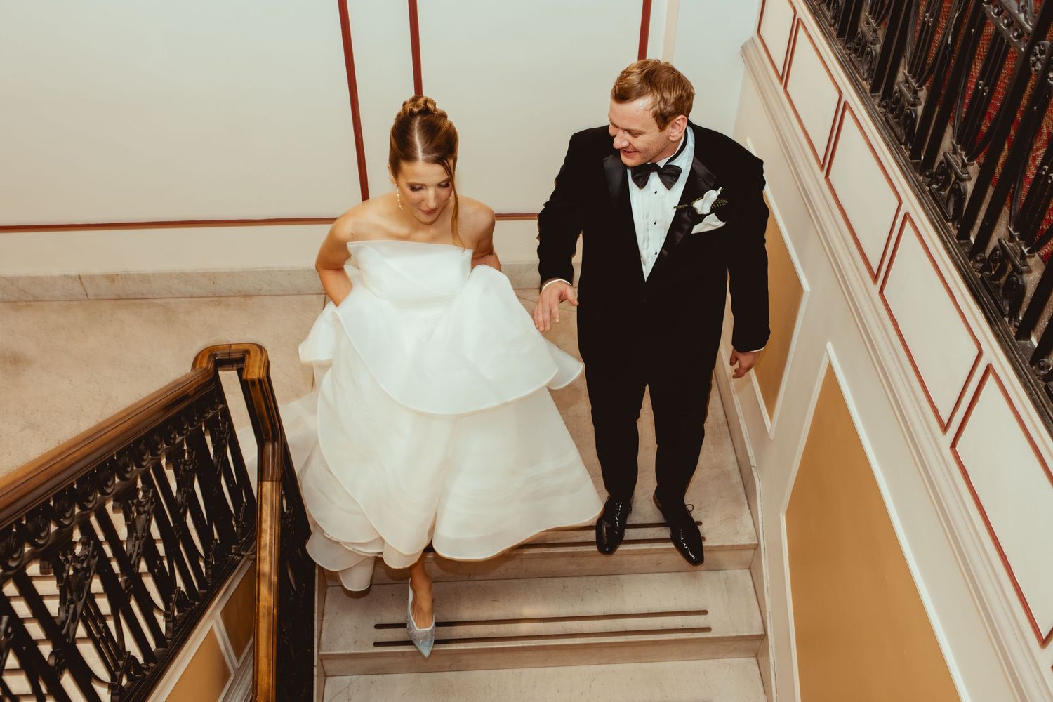 Wedding couple descends elegant staircase with ornate iron railing in a romantic indoor setting.