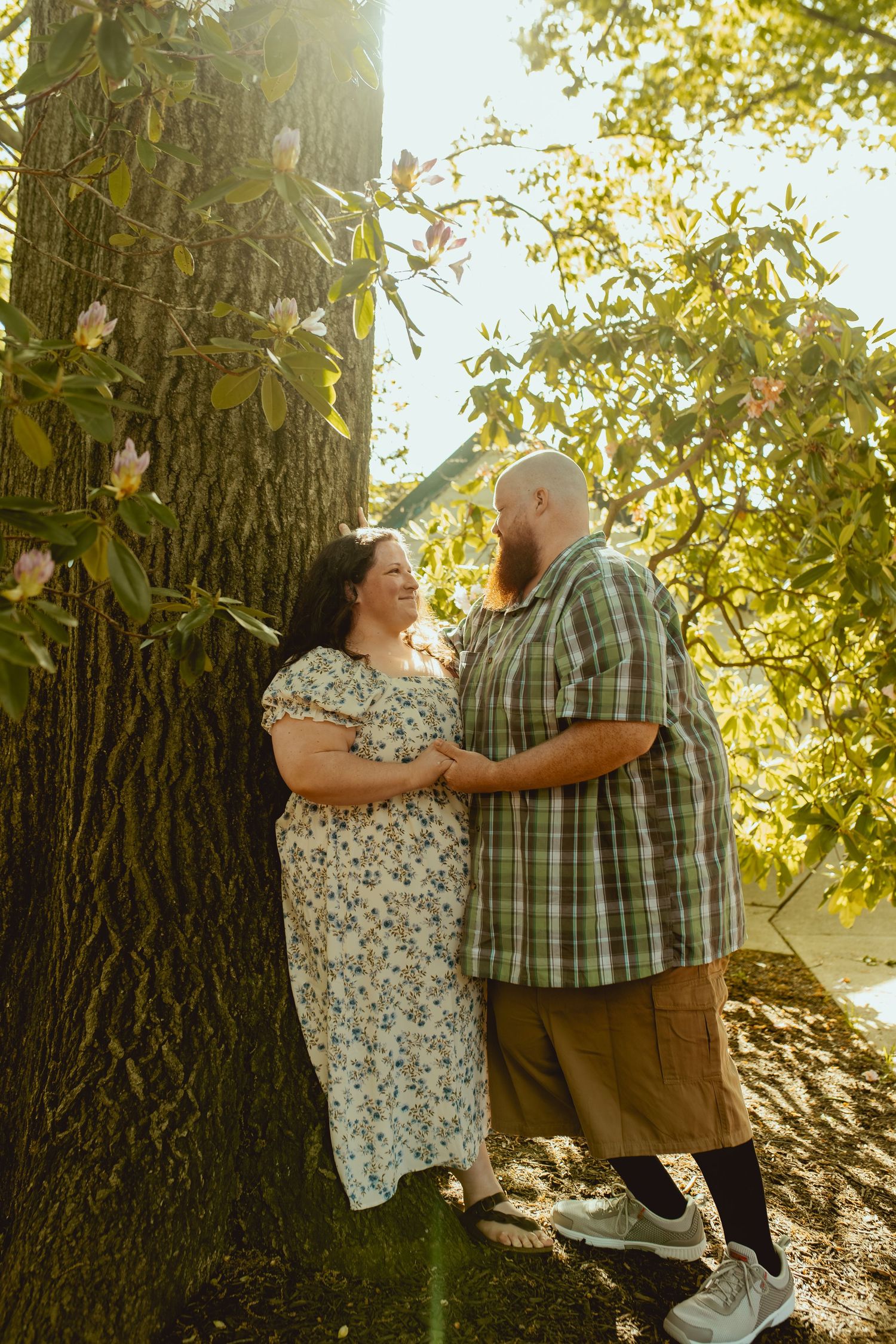 Couple holds hands and gazes at each other leaning against a large tree in a sunlit garden with blooming flowers.