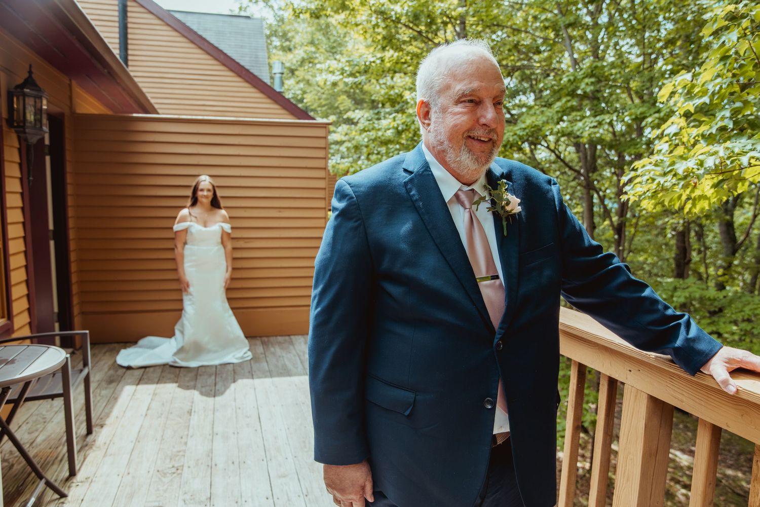 A bride in a white gown stands on a wooden deck as a man in a navy suit awaits their first look.