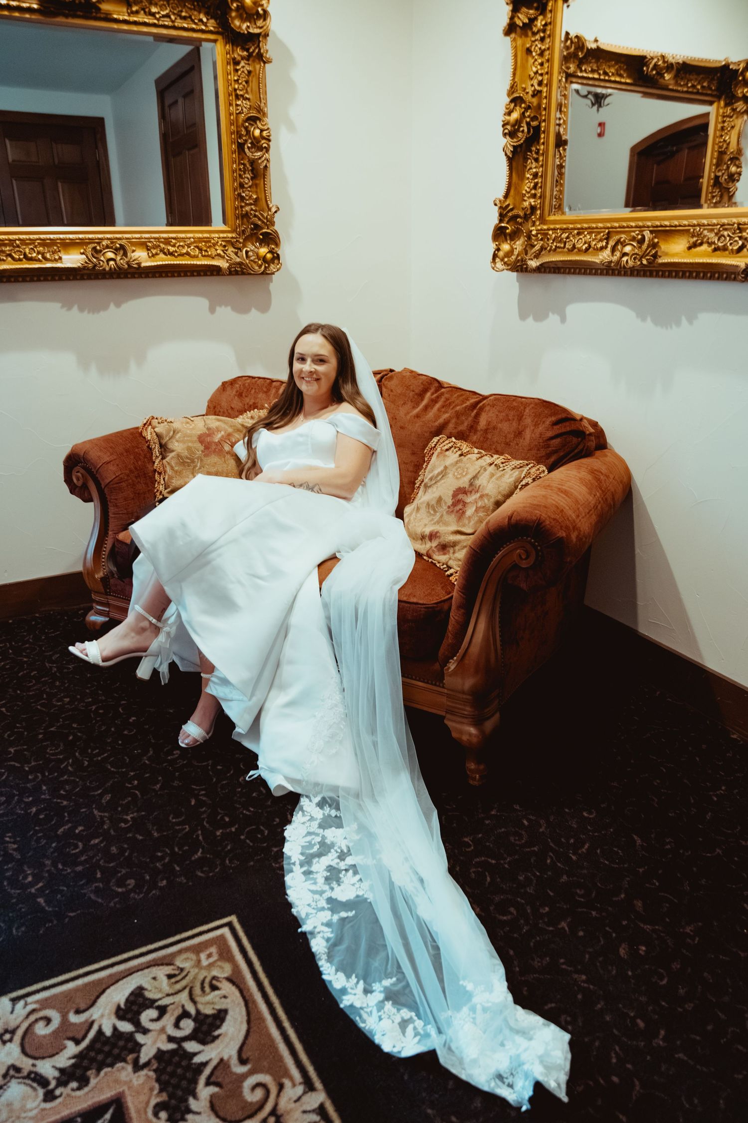 A smiling bride in a white lace wedding gown sits on a velvet sofa beneath ornate gold-framed mirrors.