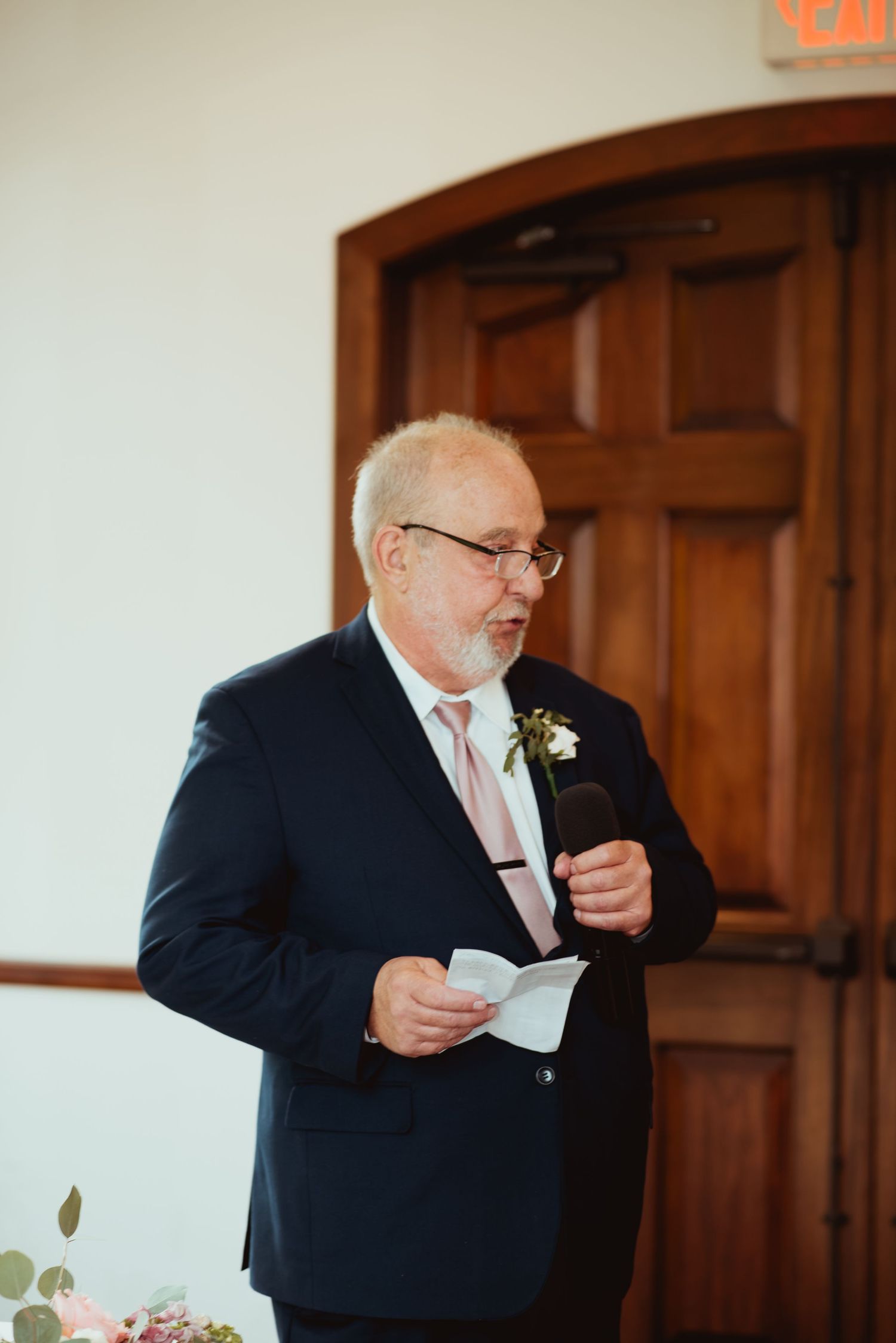 Father of the bride delivers heartfelt wedding speech holding notes in navy suit with boutonniere.