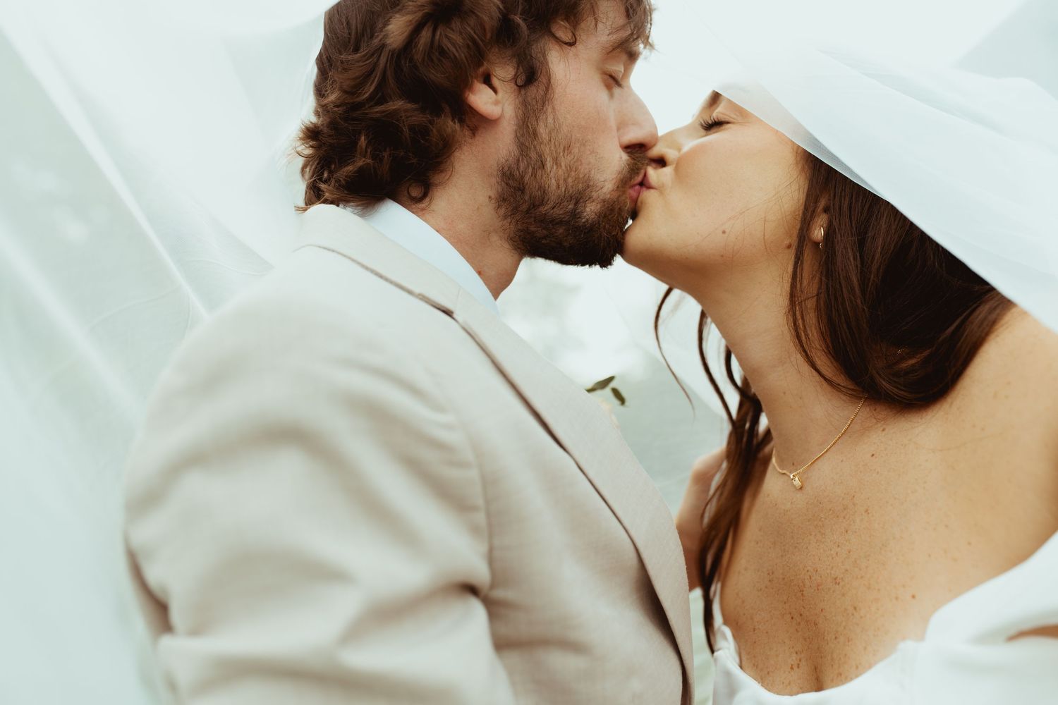 A bride and groom share a romantic kiss, with the bride wearing a white veil and the groom in a white suit.