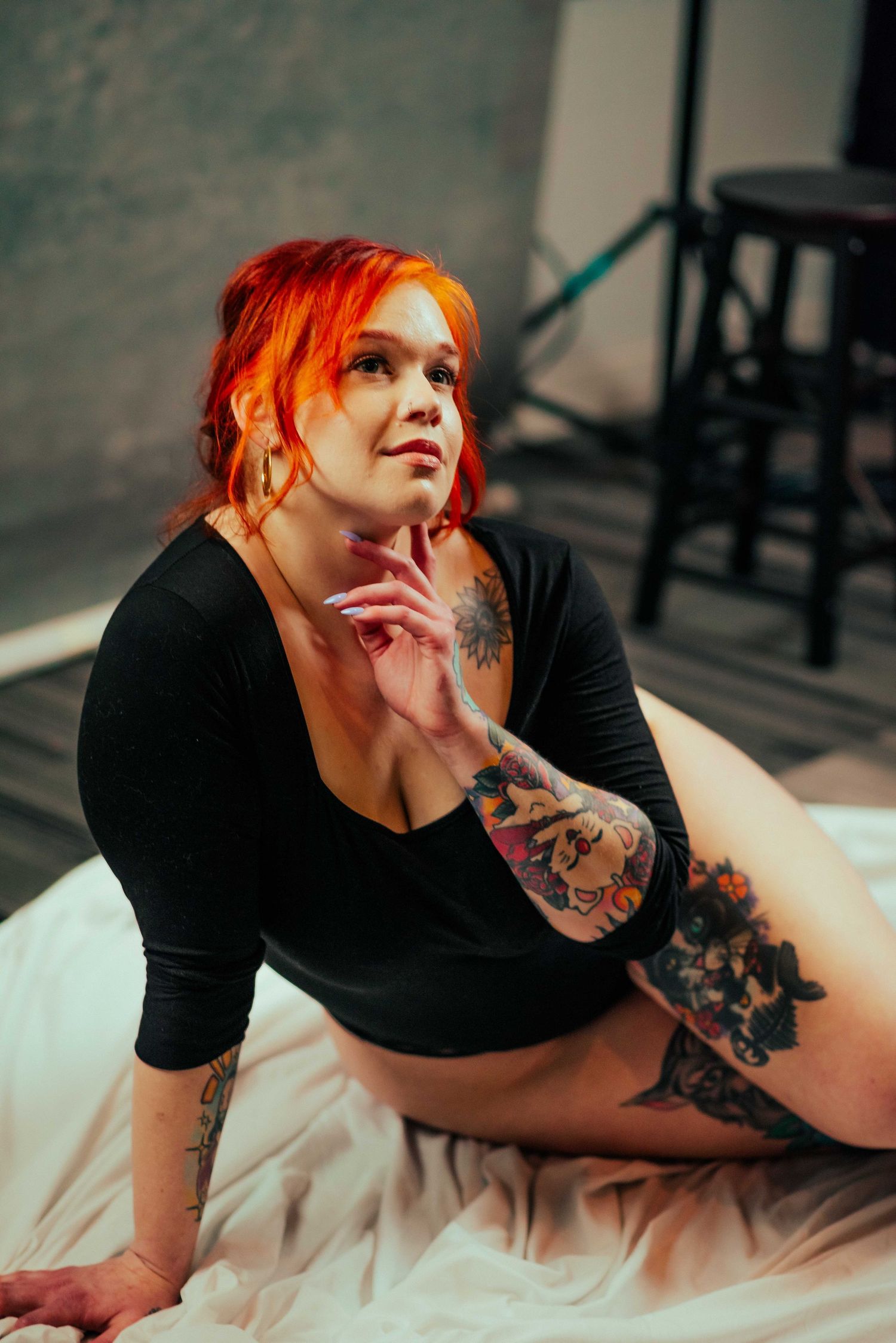 A plus-size woman with vibrant red hair and tattoos poses confidently in a black outfit on a white sheet in a studio.