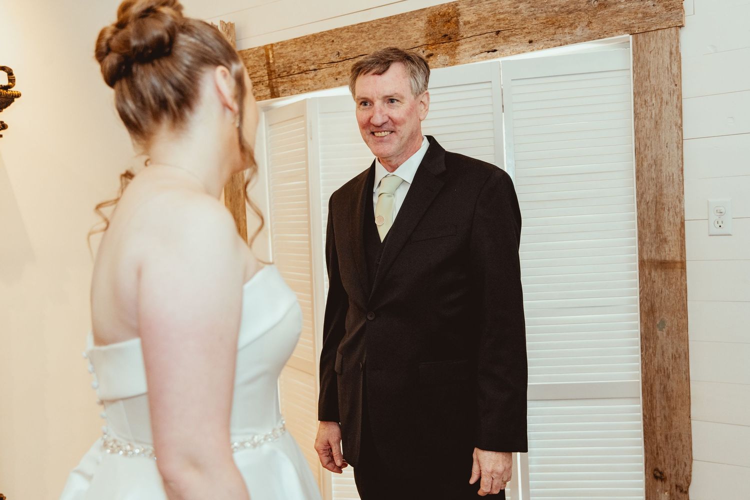 A proud father smiles warmly at his daughter the bride in her elegant white strapless wedding gown indoors.