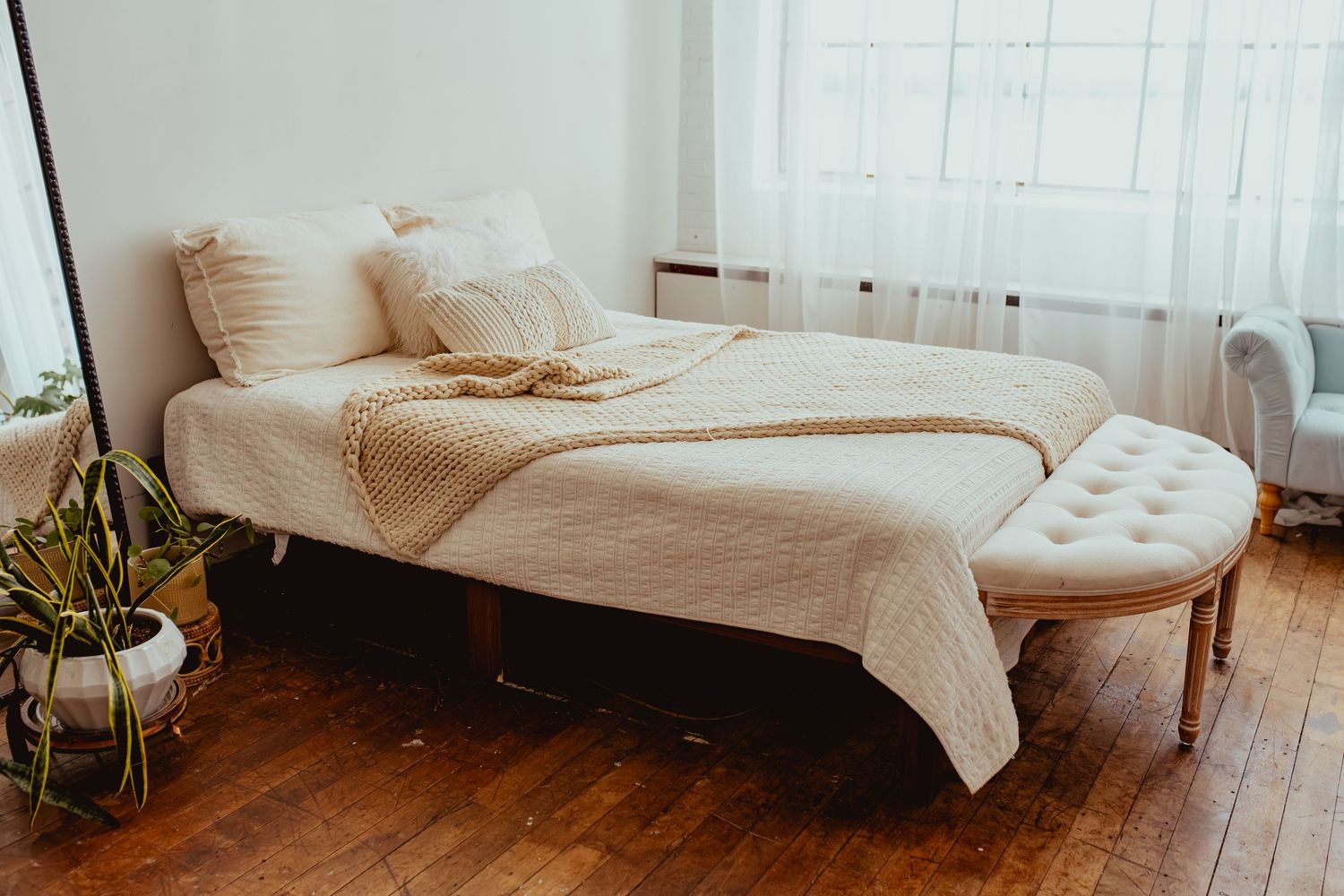 Cozy bedroom with a white bed, knit throw blanket, tufted bench, hardwood floors, and a bright window.