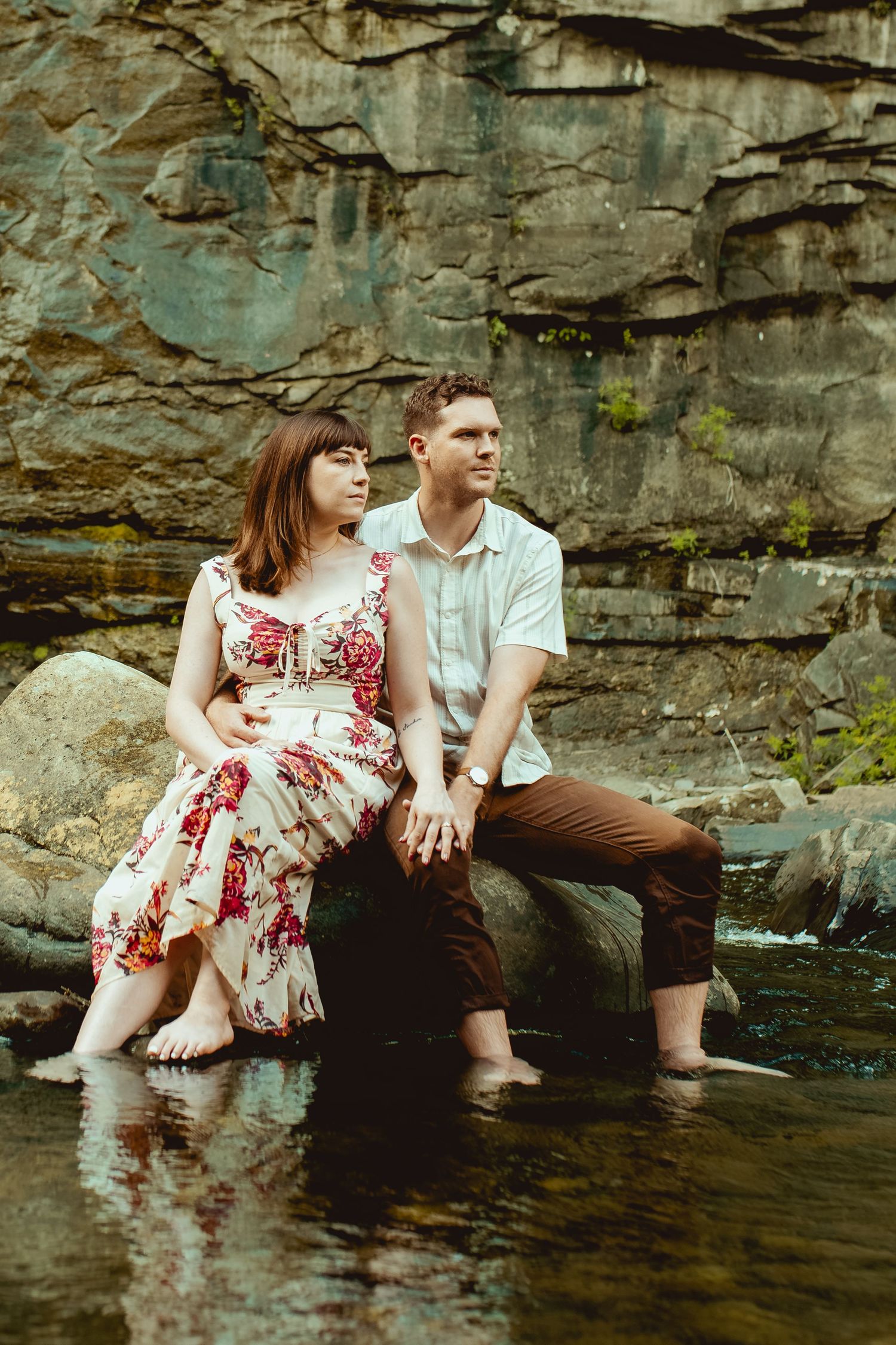Couple sitting barefoot on rocks by a stream with a stone cliff backdrop, woman in floral dress, man in casual attire.