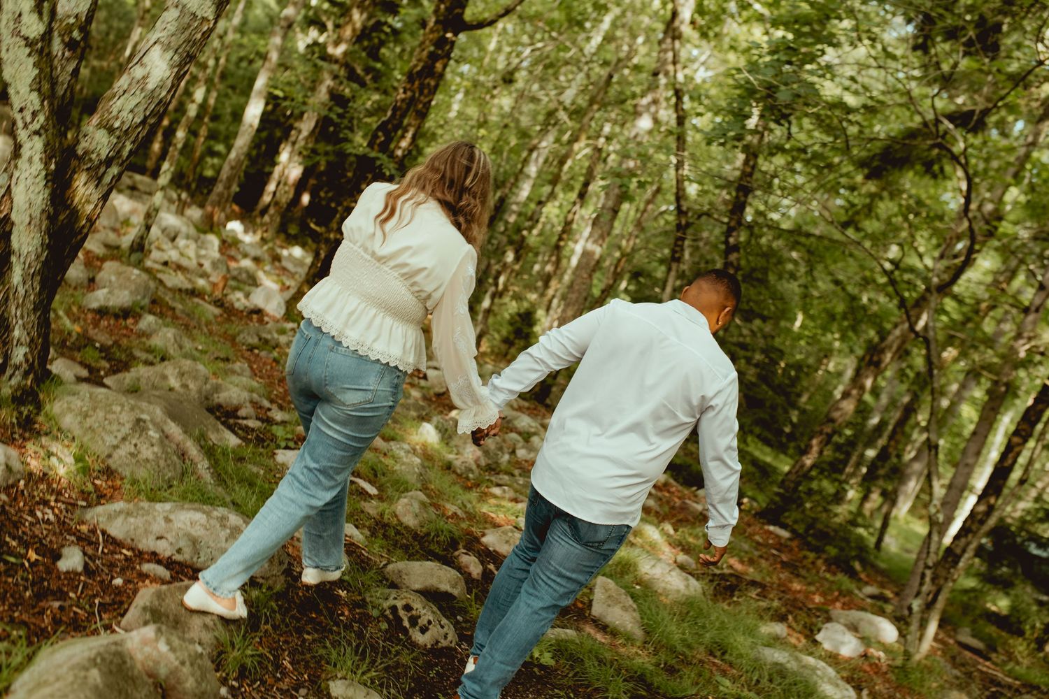 Couple holding hands hiking through a lush green forest on rocky terrain.