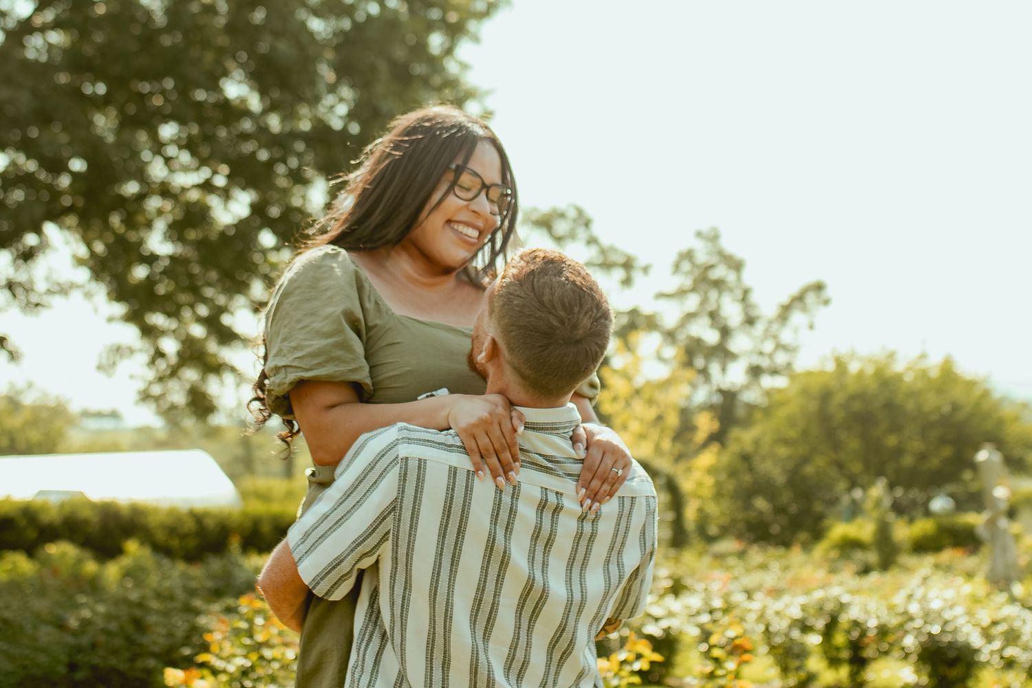 A man lifts a smiling woman in a sunny garden, sharing a joyful romantic moment outdoors.