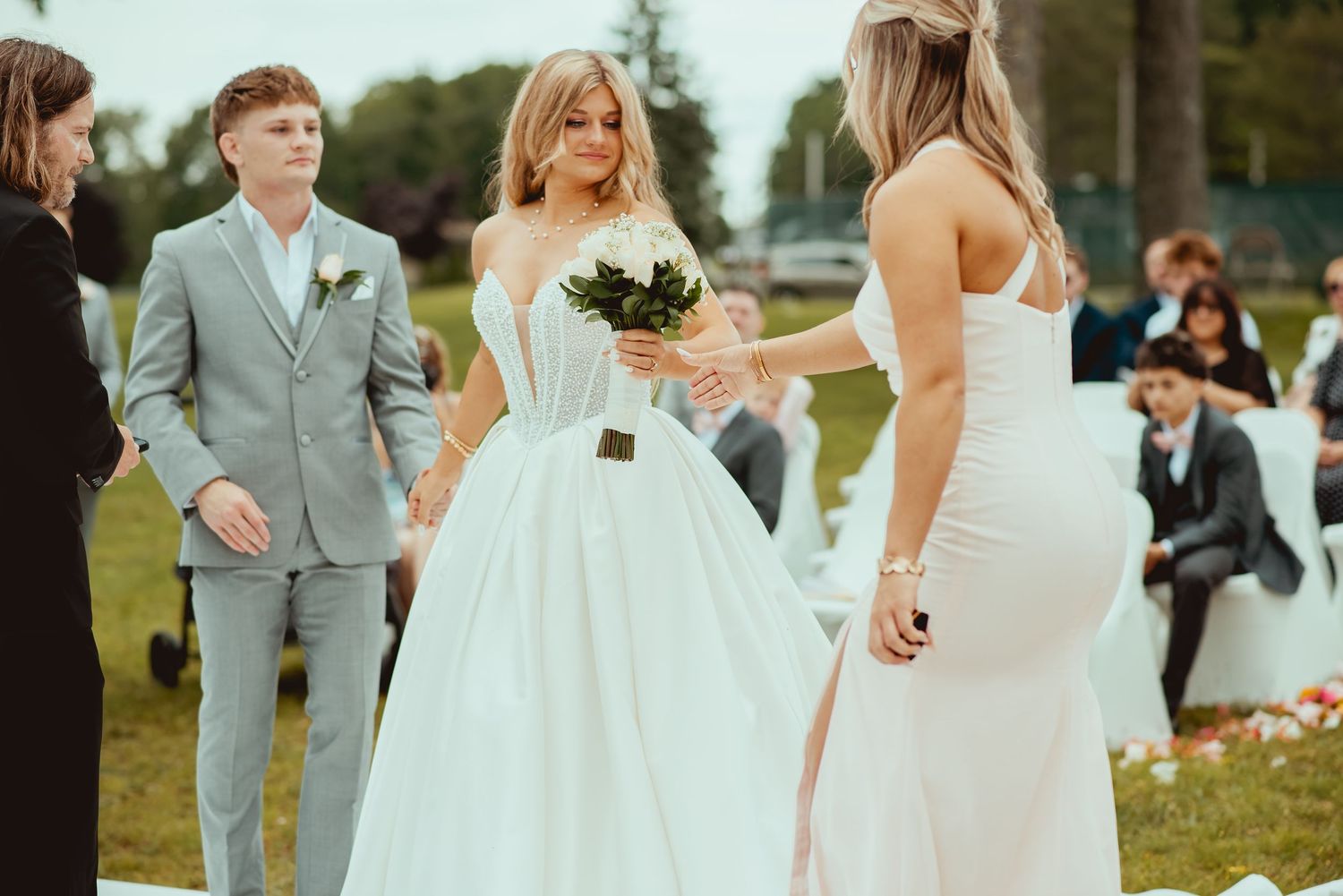 Bride in white ball gown receives bouquet from bridesmaid at outdoor wedding ceremony.
