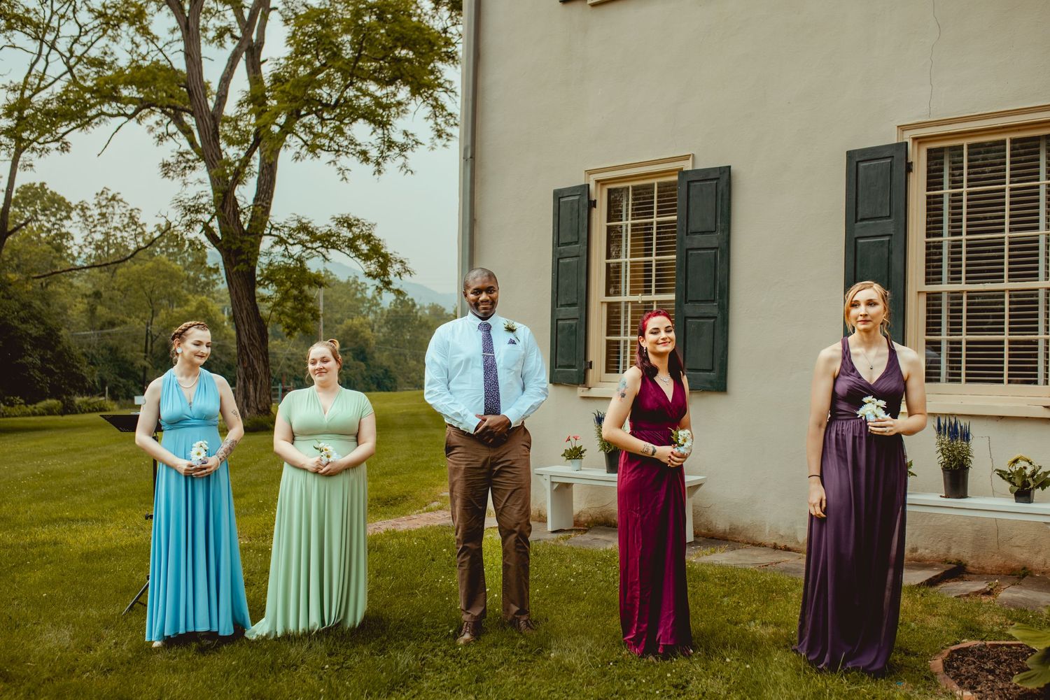 Wedding party members stand in a row on a lawn outside a historic white house with green shutters.