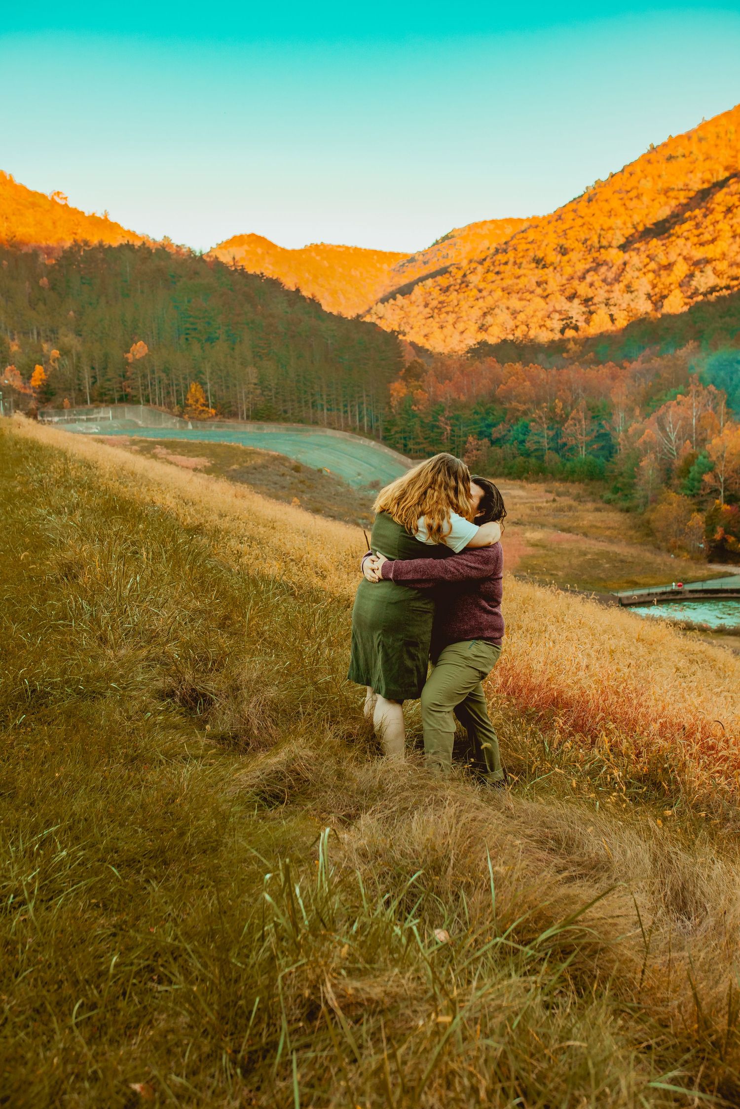 A couple embraces in a warm moment while standing on a grassy hillside overlooking a scenic autumn valley at sunset.