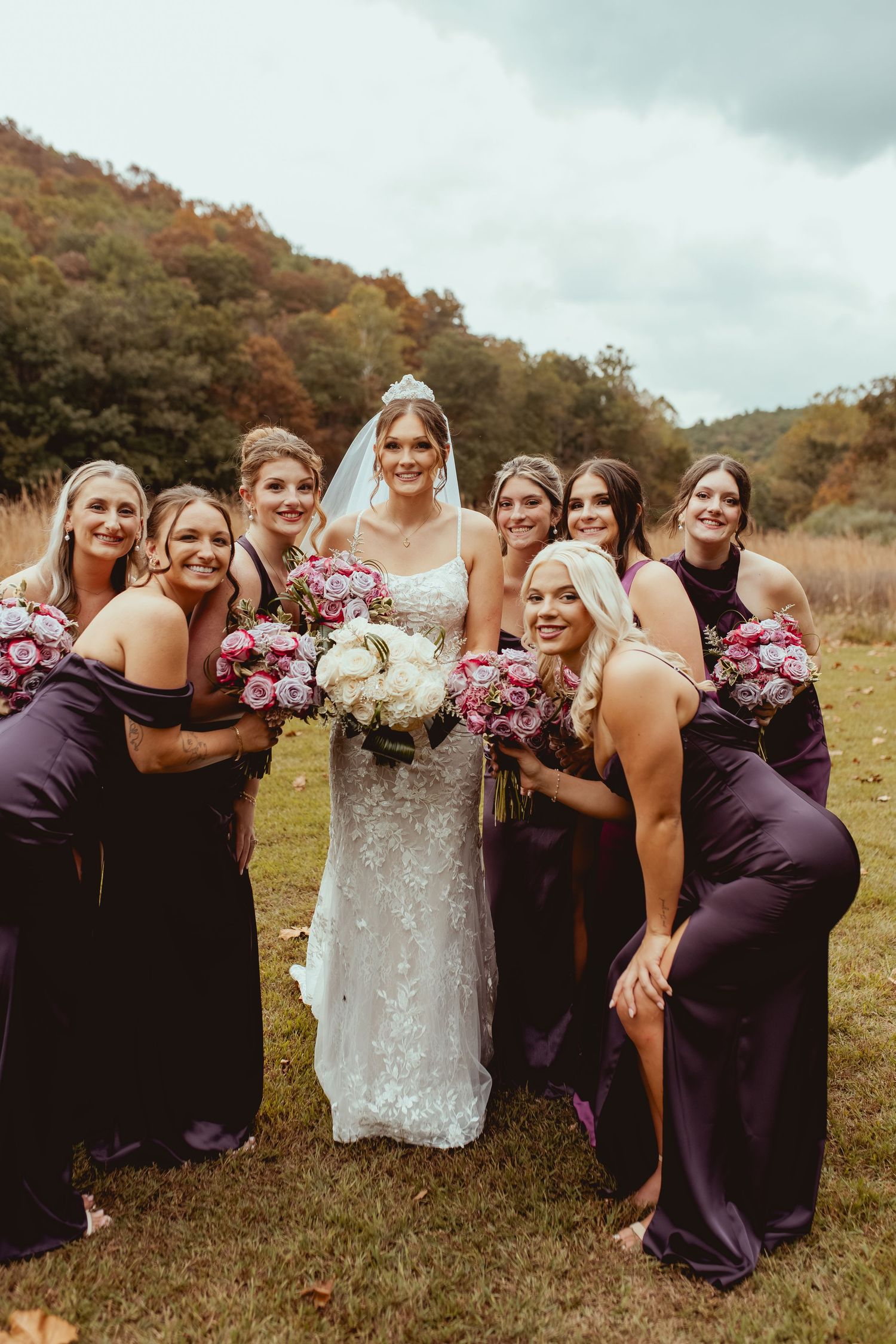 Bridesmaids in purple dresses pose with bouquets against a scenic hillside backdrop.