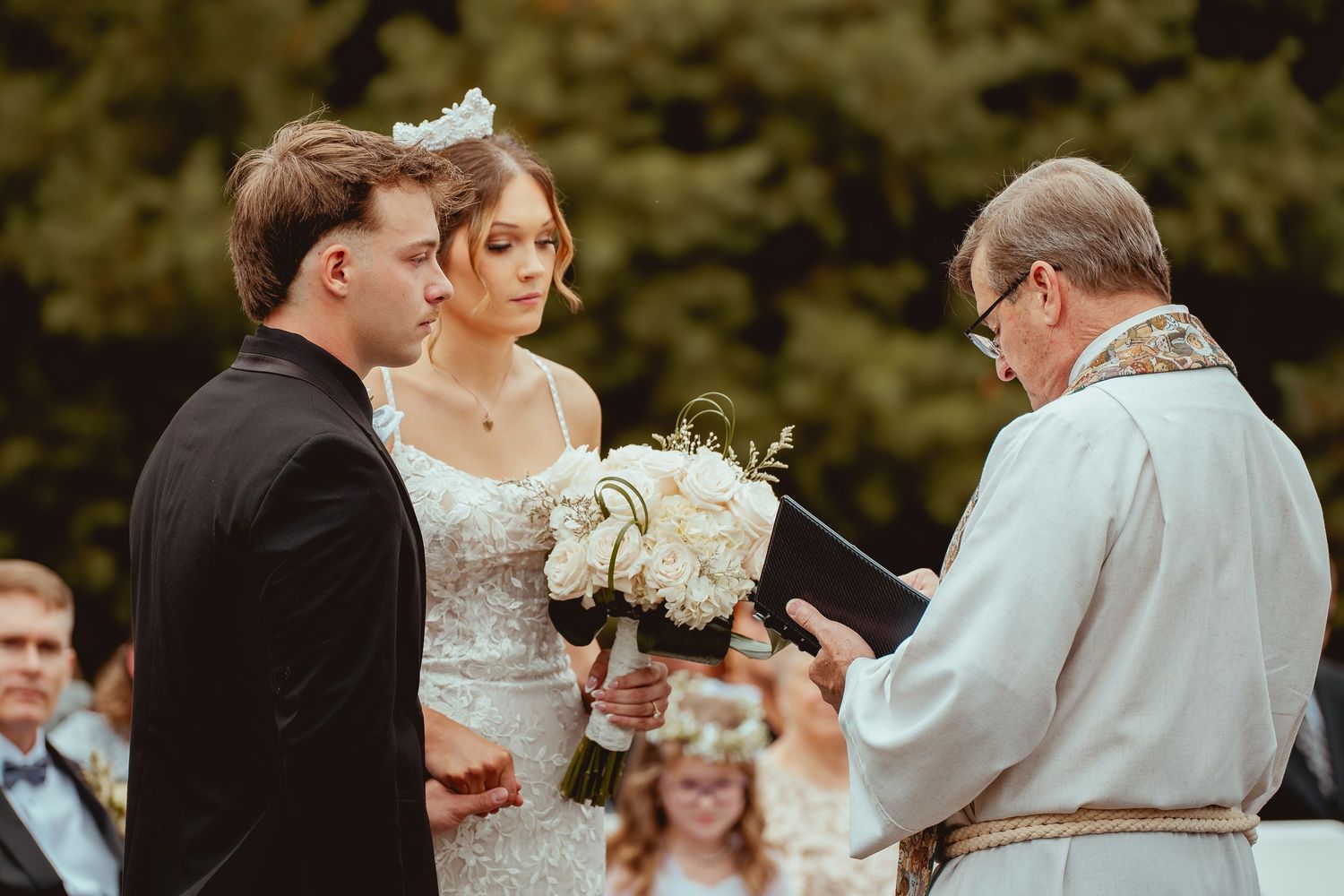 A wedding ceremony takes place on a hillside with guests seated in white chairs.