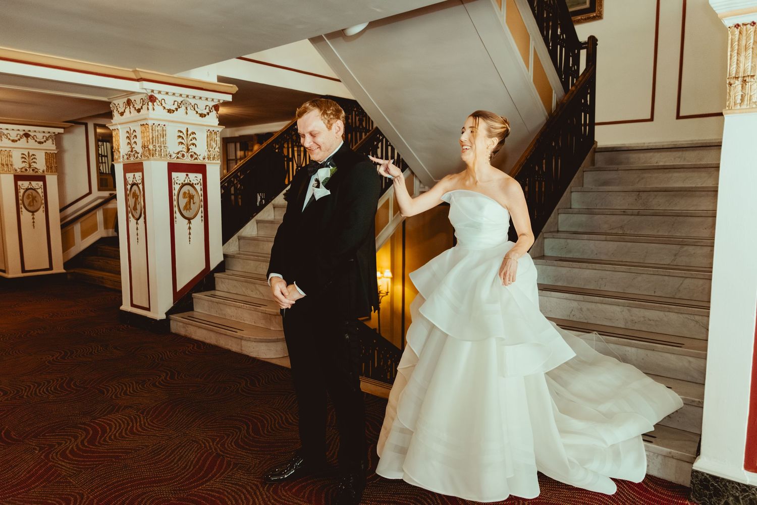Bride in white gown descends elegant staircase in historic building with ornate details.