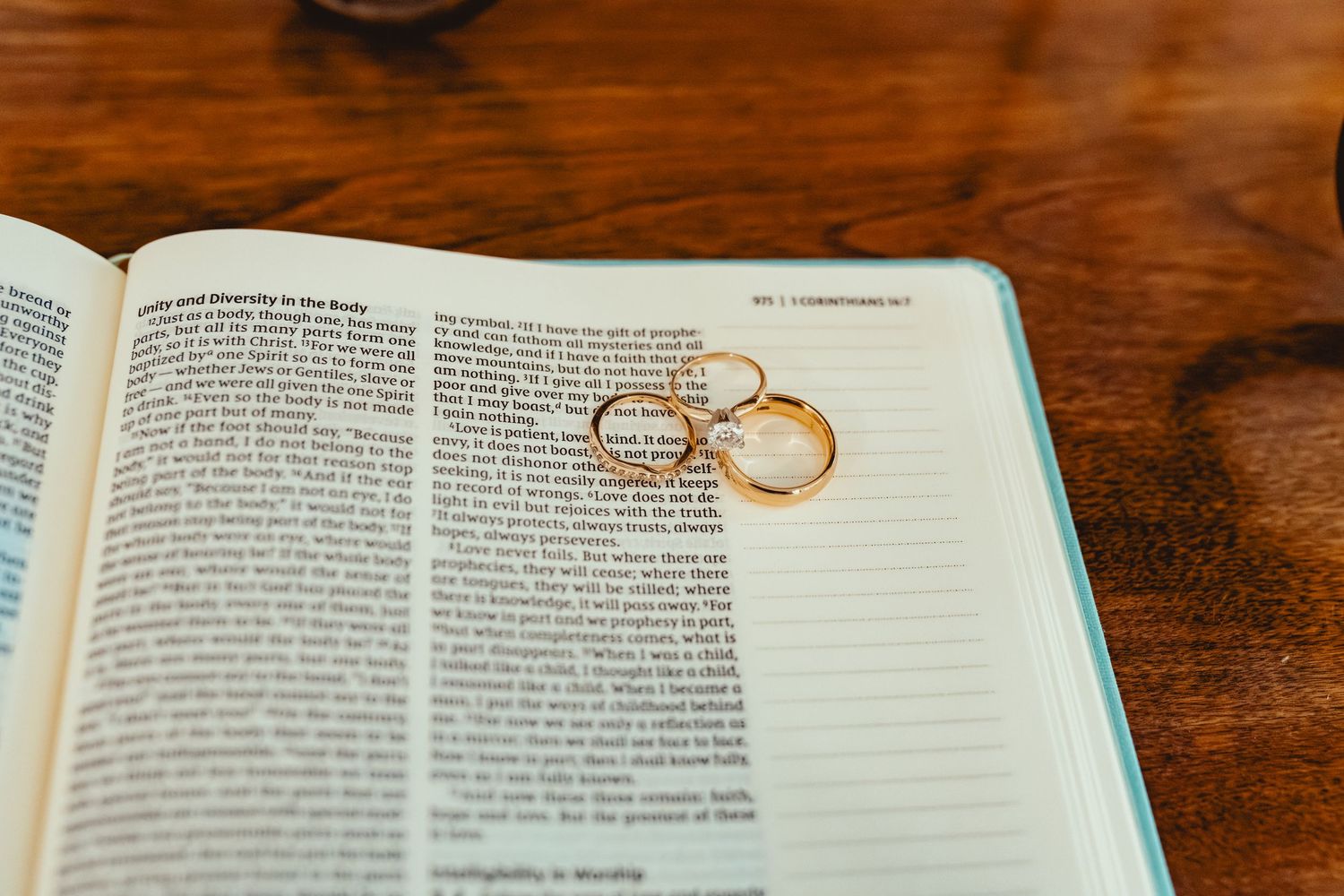 Wedding rings rest on an open Bible page on a wooden surface in natural lighting.