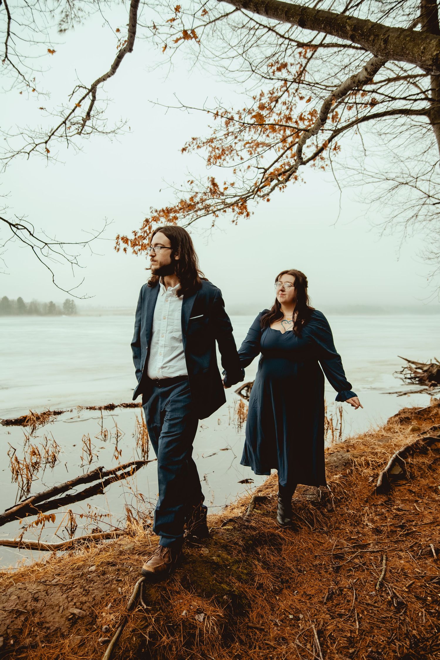 Two people stand on an autumn shoreline overlooking a misty lake with bare branches overhead.