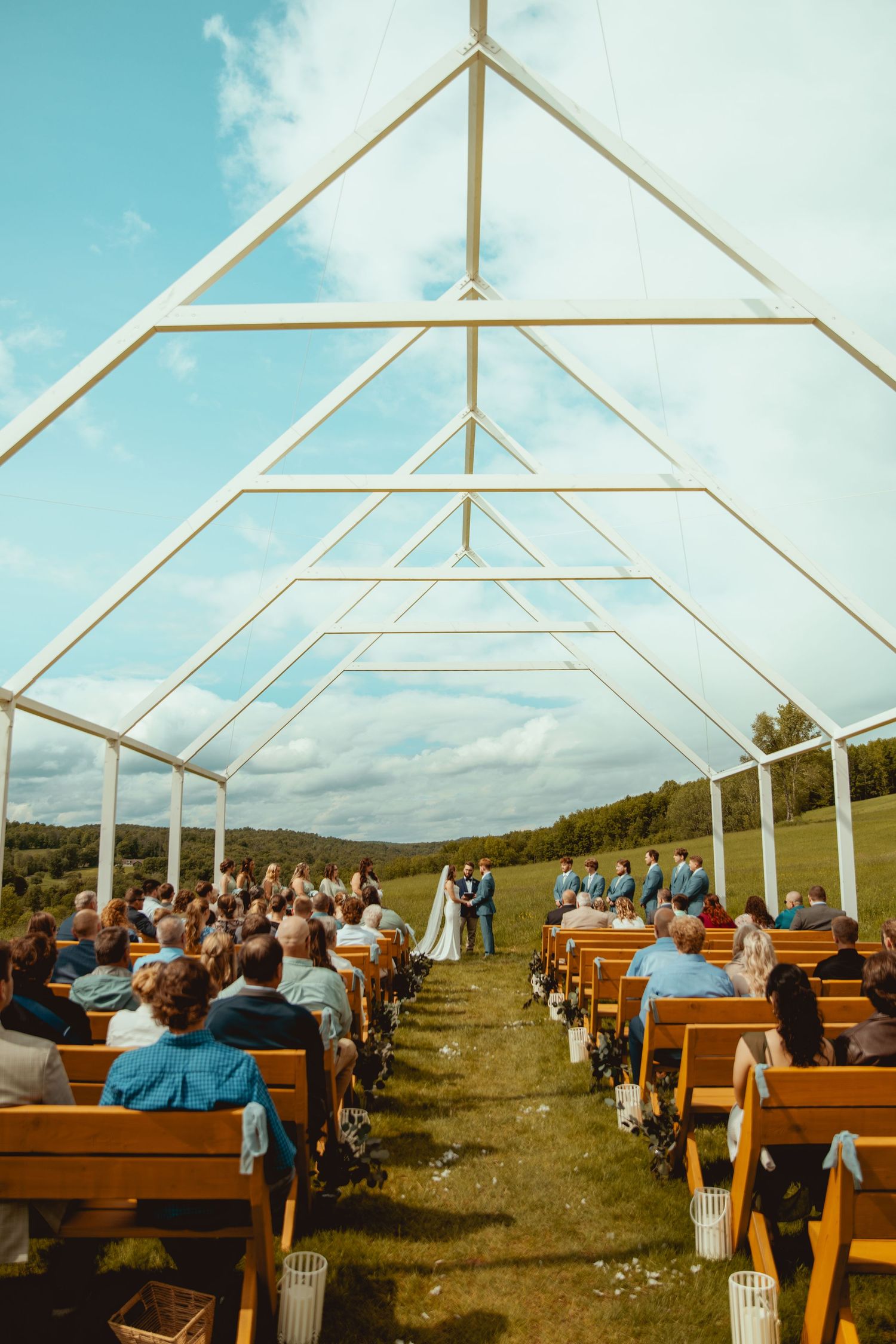 Outdoor wedding ceremony setup with white tent frame and wooden pews against mountain backdrop.