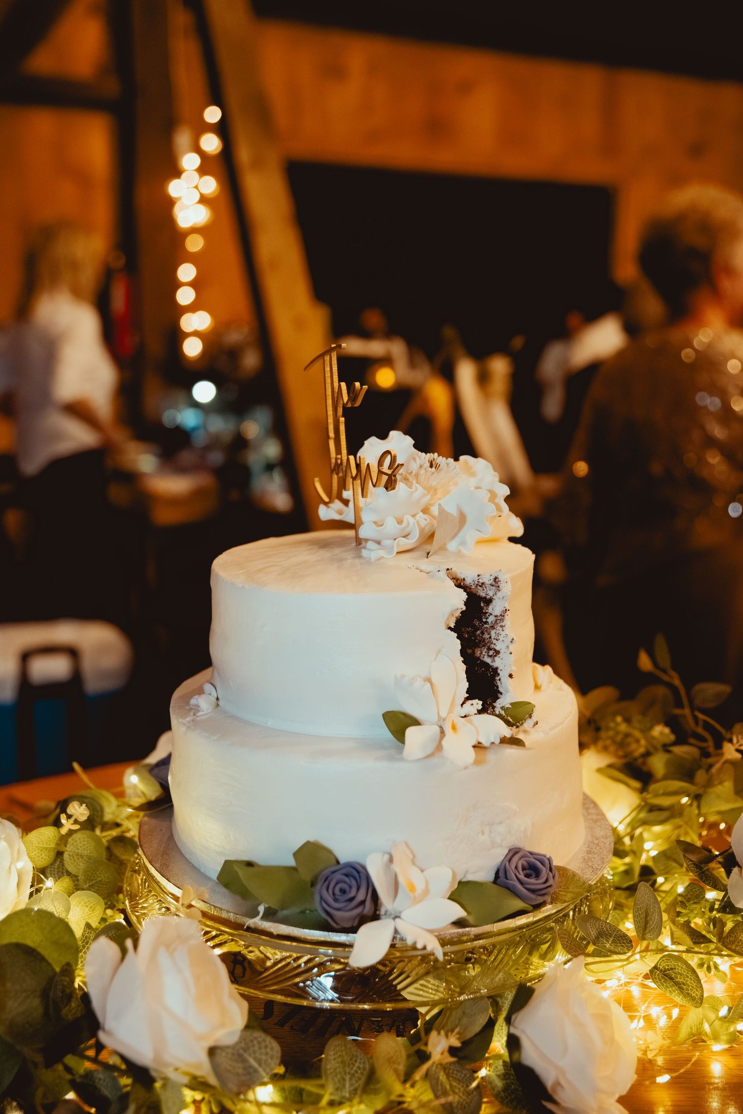 A white tiered wedding cake decorated with purple flowers and greenery sits illuminated by warm string lights at a reception.