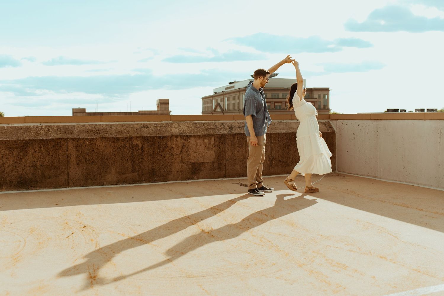 Couple dancing on a rooftop terrace, man spinning woman in white dress under a blue sky.