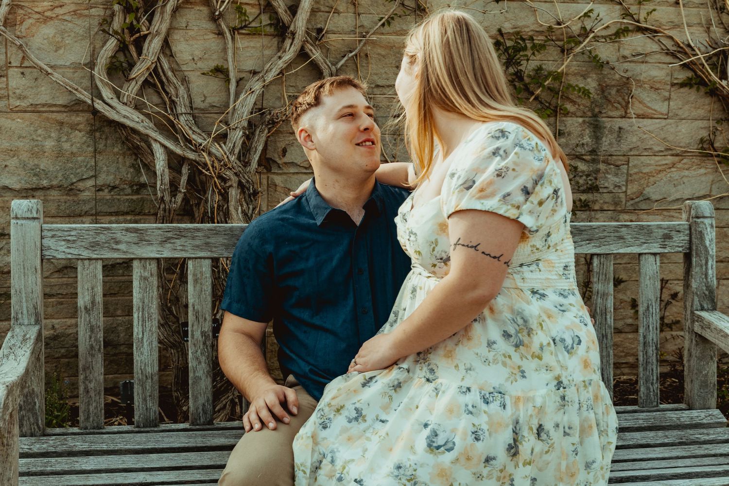 Couple sitting on a wooden bench outdoors, woman in floral dress and man in blue shirt sharing a romantic moment.