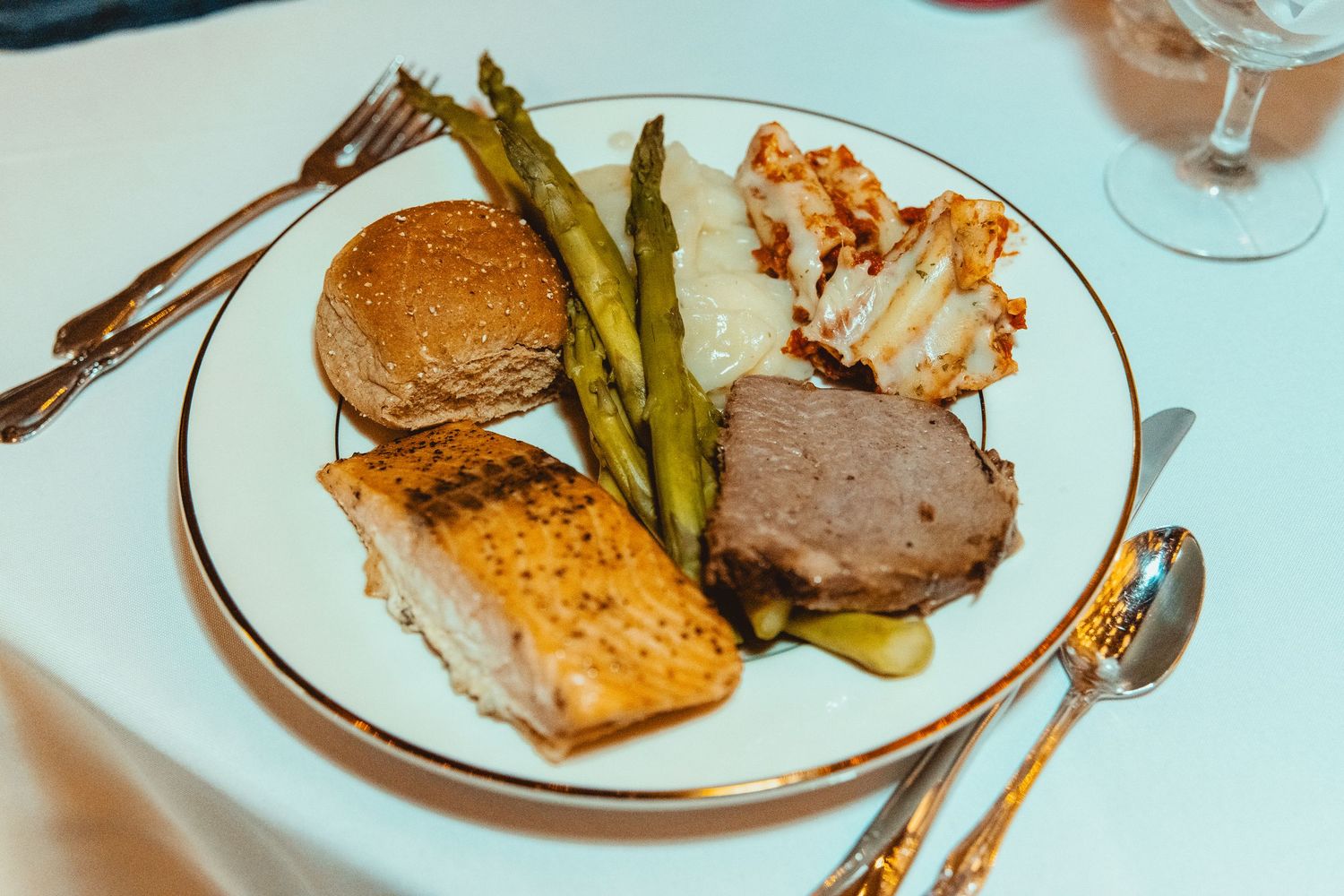 Elegant wedding reception dinner plate with salmon, asparagus, meat, and a roll on white gold-rimmed china.