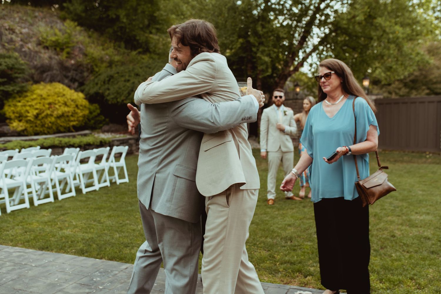 Two men share a warm hug at an outdoor wedding ceremony while a woman in blue looks on smiling.