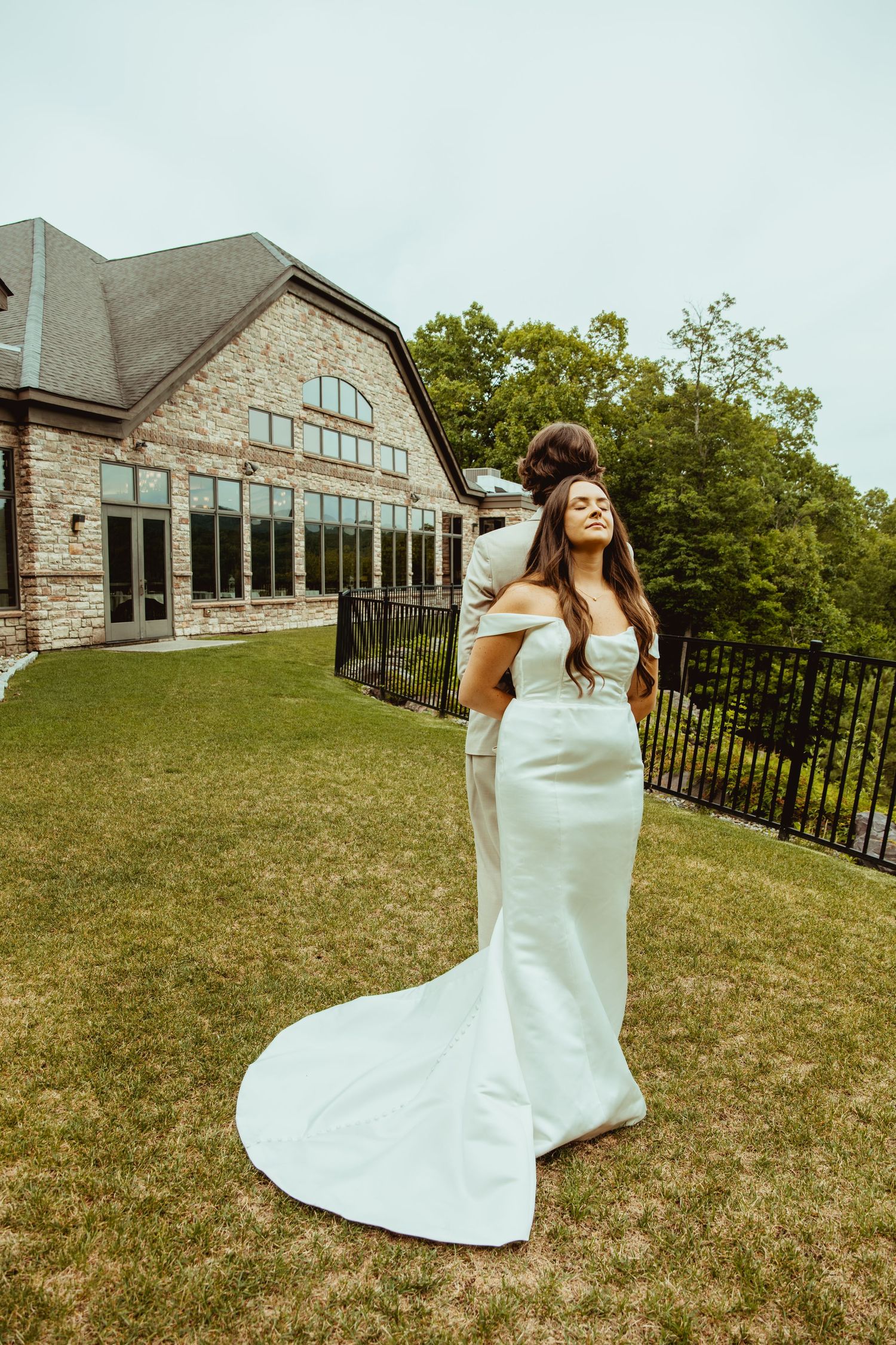 Bride in elegant white off-shoulder gown with train stands on lawn outside stone venue, groom embracing her from behind.