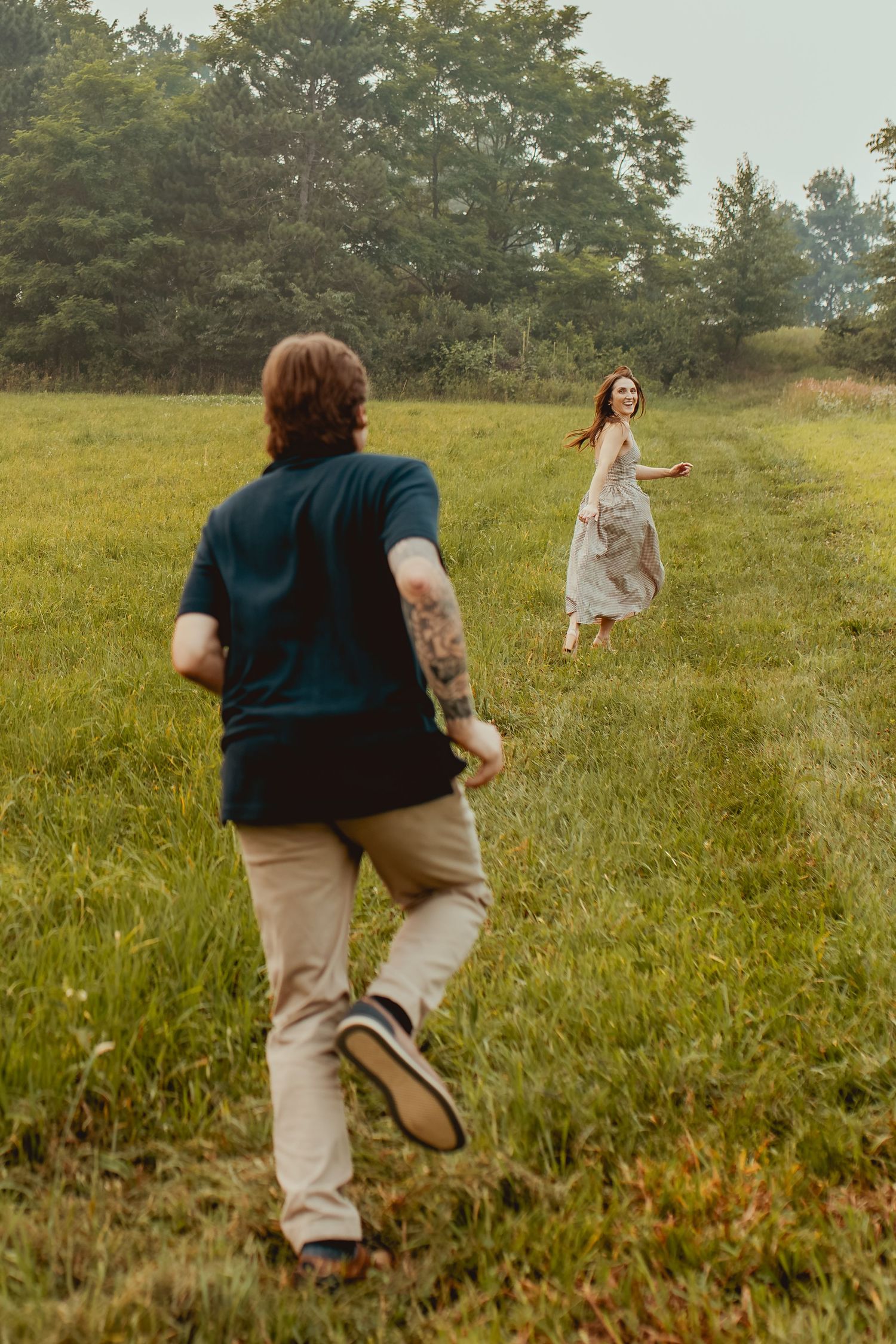 A tattooed man chases a laughing woman in a white dress through a lush green field with trees in the background.