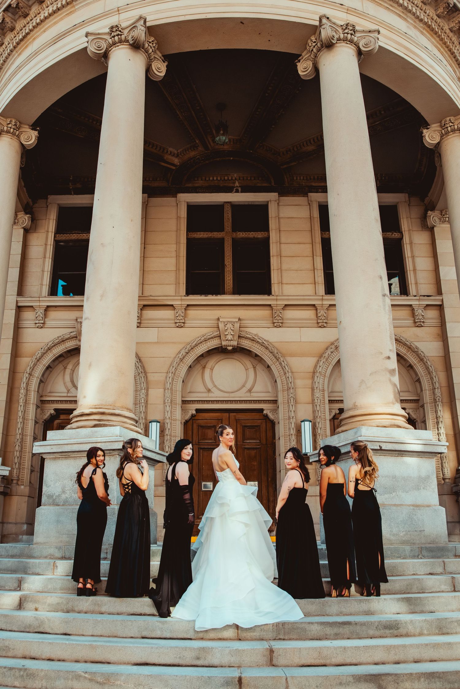 Wedding party poses on grand stone steps with classical columns in the background.