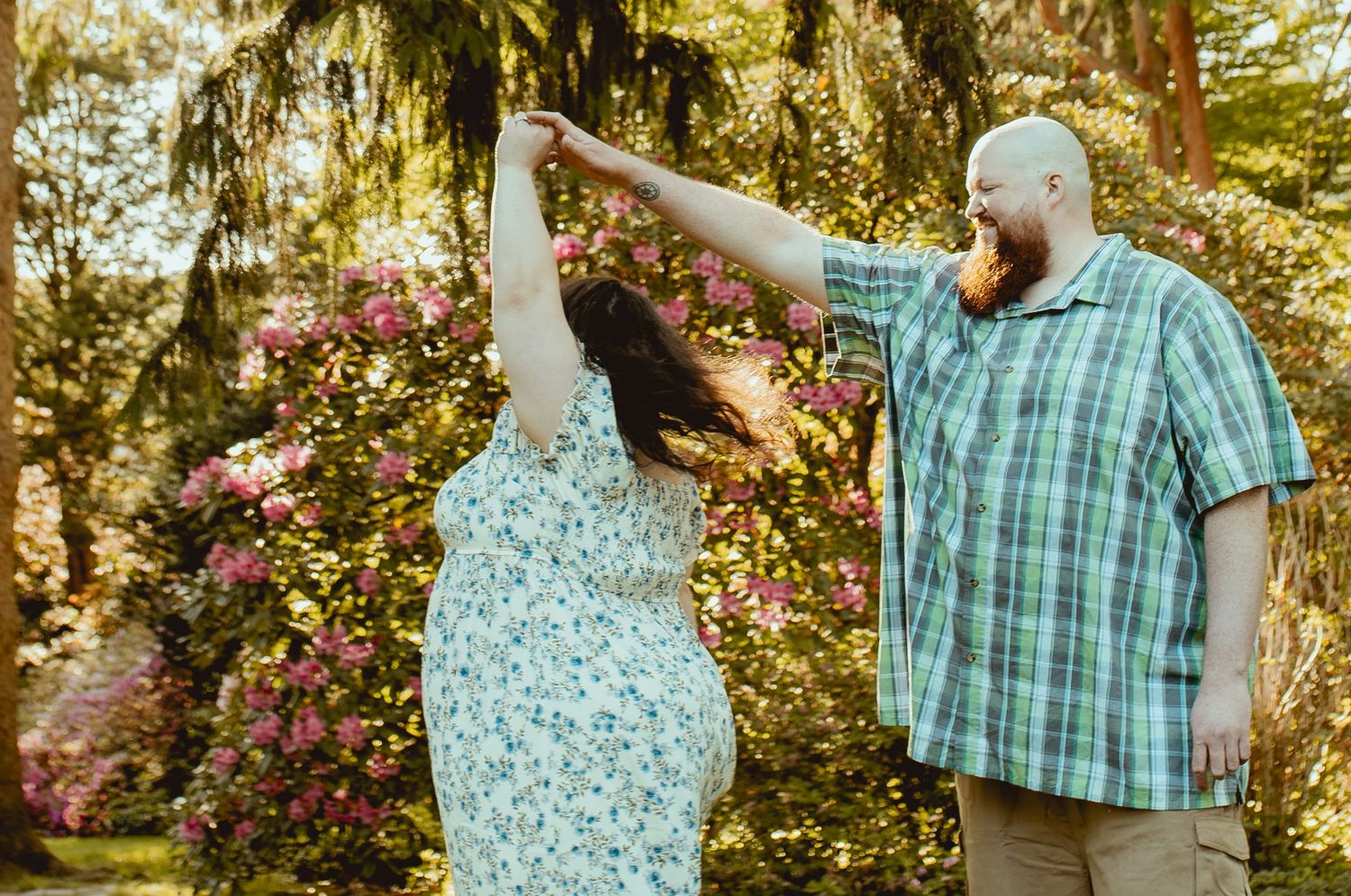A couple dancing outdoors among blooming pink flowers, man in plaid shirt spinning woman in floral dress.