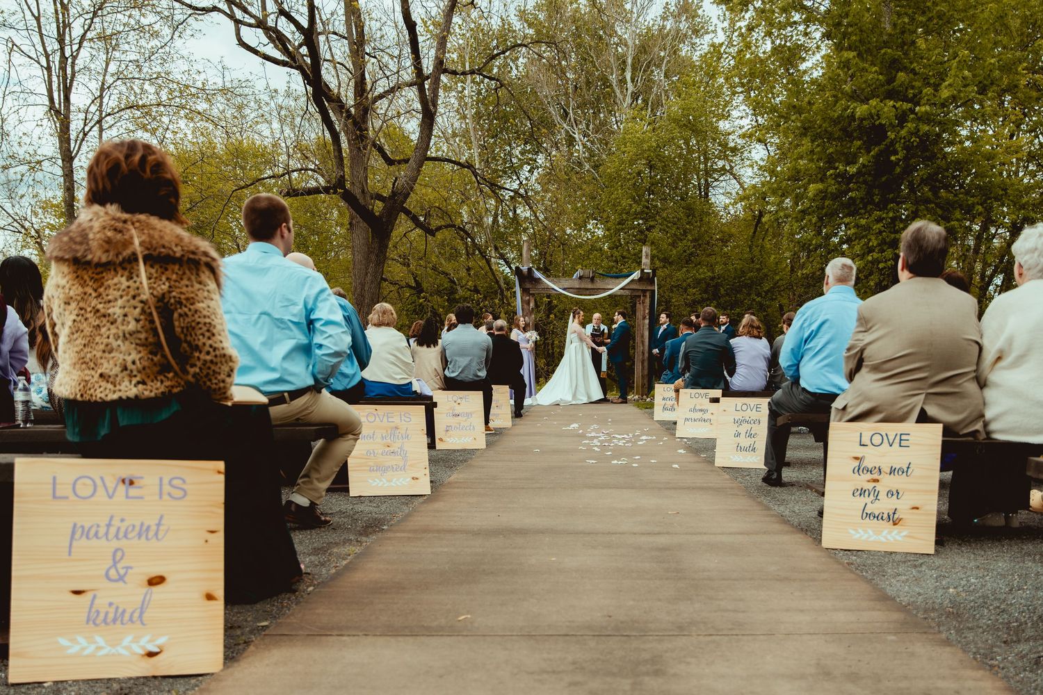 Outdoor wedding ceremony with guests seated on benches lined with rustic love quote signs, couple at floral arch.