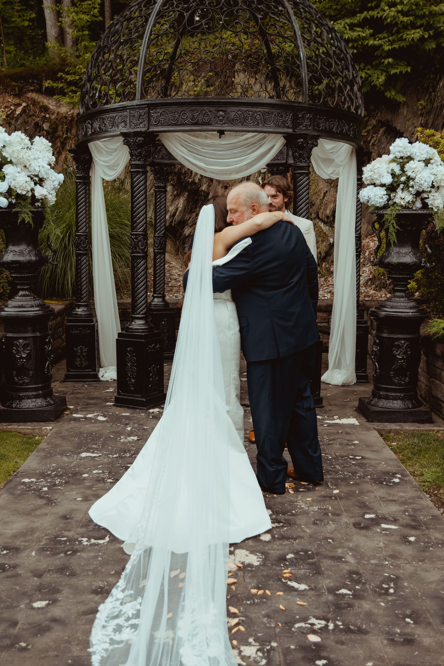 Bride and groom share a tender embrace under ornate gazebo with white floral arrangements.