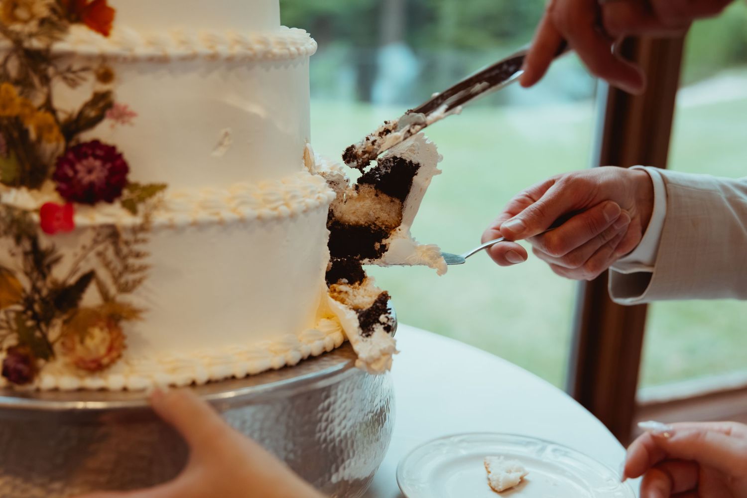 Newlyweds cut into a elegant white tiered wedding cake with floral decorations revealing a dark filling.